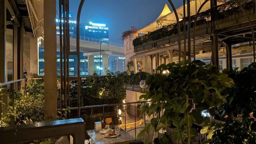 Nighttime scene of an outdoor restaurant or café with tables set for dining. The area is decorated with lush greenery and string lights, with a cityscape of illuminated buildings and signs visible in the background.