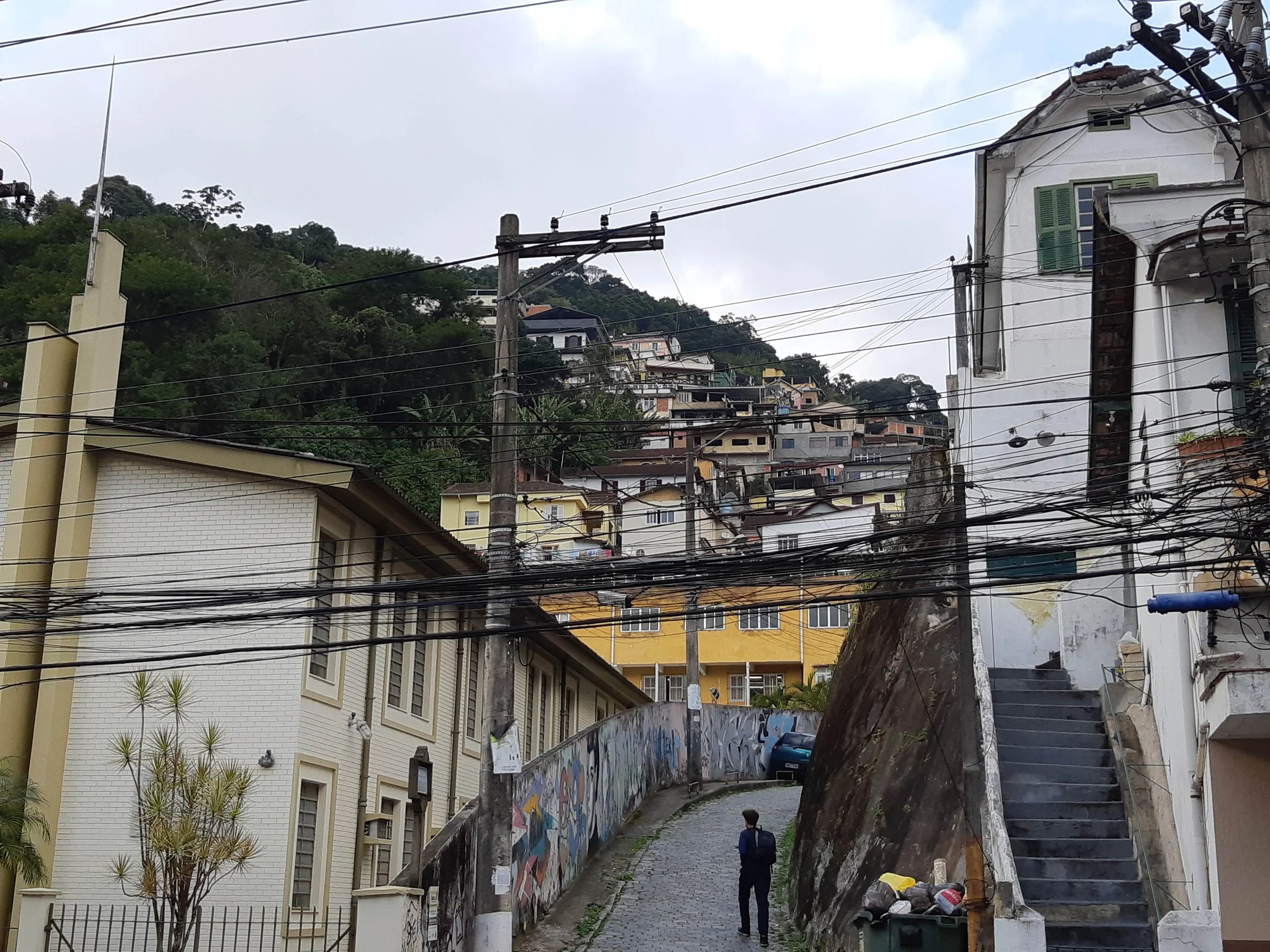 Hilly street scene with multiple residential buildings, electrical wires overhead, and a person walking on the cobblestone sidewalk.