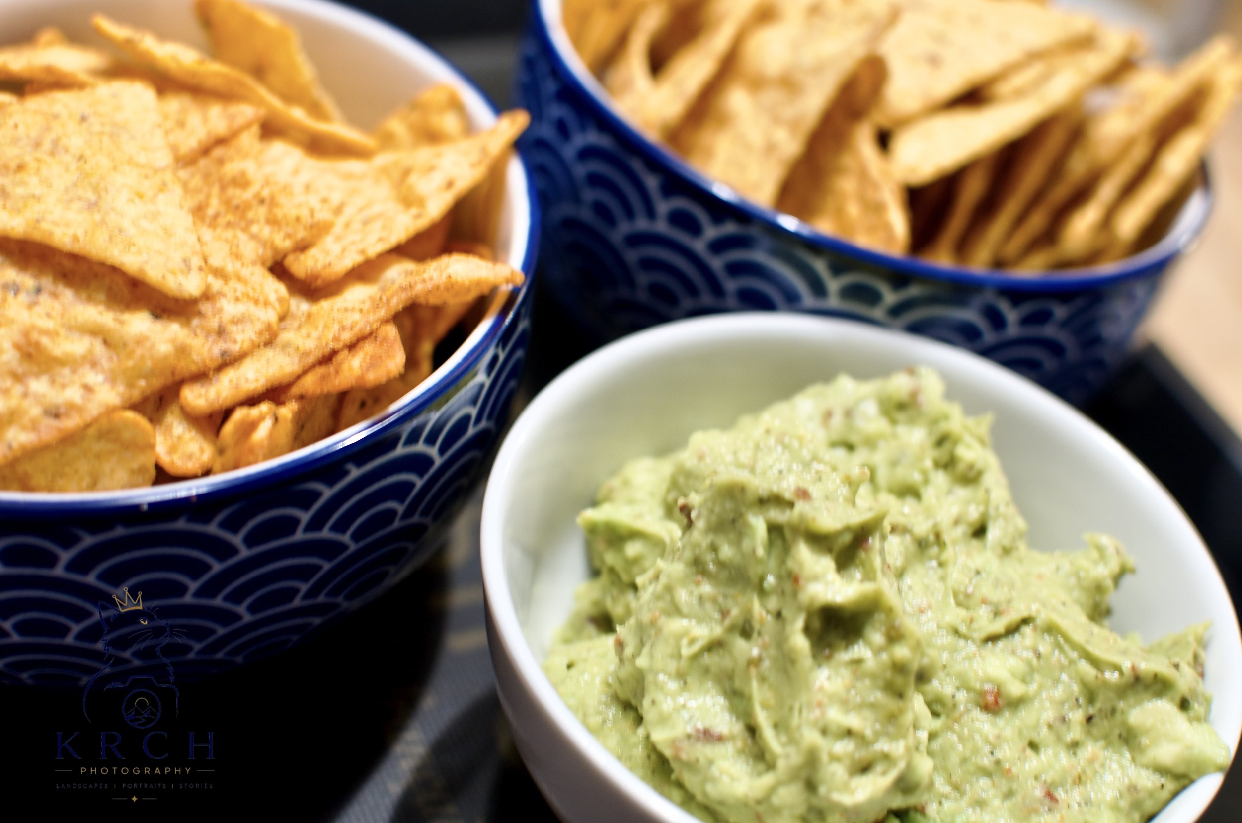 Close-up of two bowls filled with tortilla chips and guacamole, with one bowl containing orange chips and the other containing yellow chips, and a small white bowl of guacamole in the foreground.