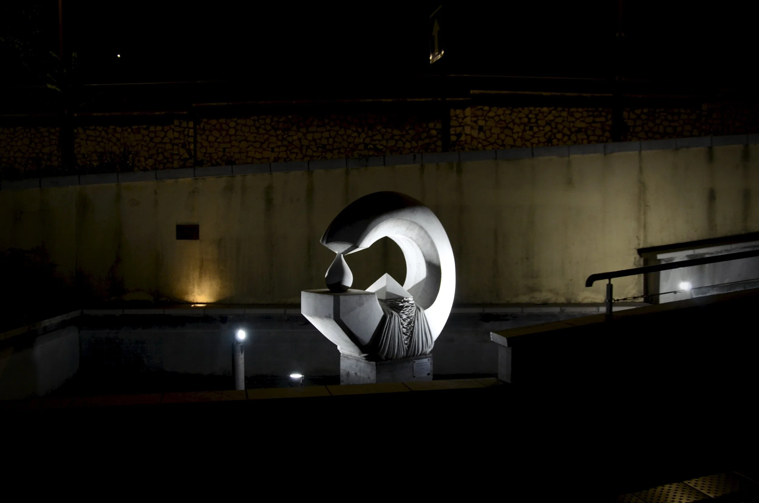 Nighttime photo of a modern abstract sculpture with curved metallic elements on a pedestal, illuminated by lights. Background includes a concrete wall, street railing, and a stacked wood fence.