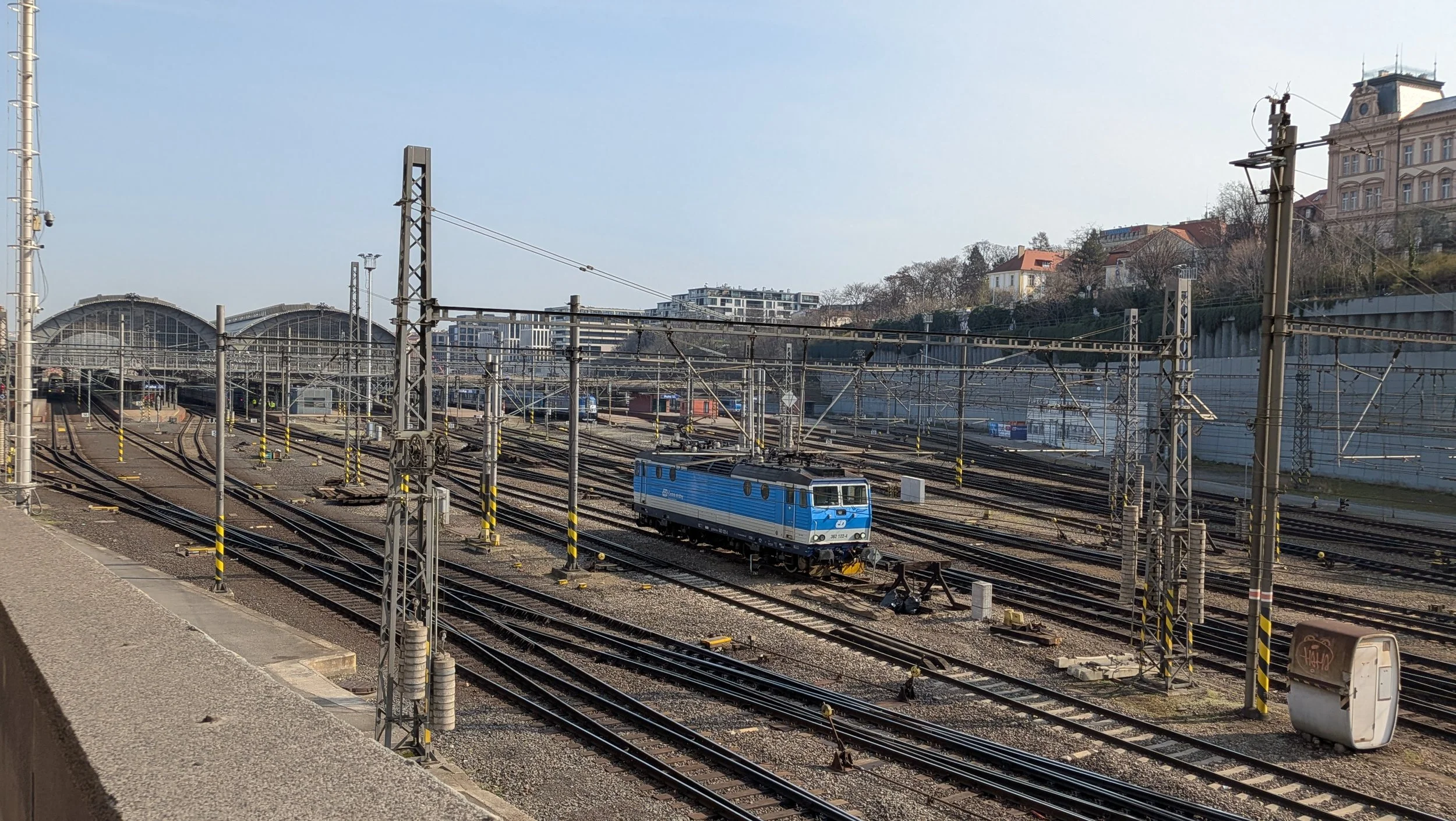 A train station with multiple train tracks, an electric locomotive, and overhead power lines against buildings and a hill with houses and trees.