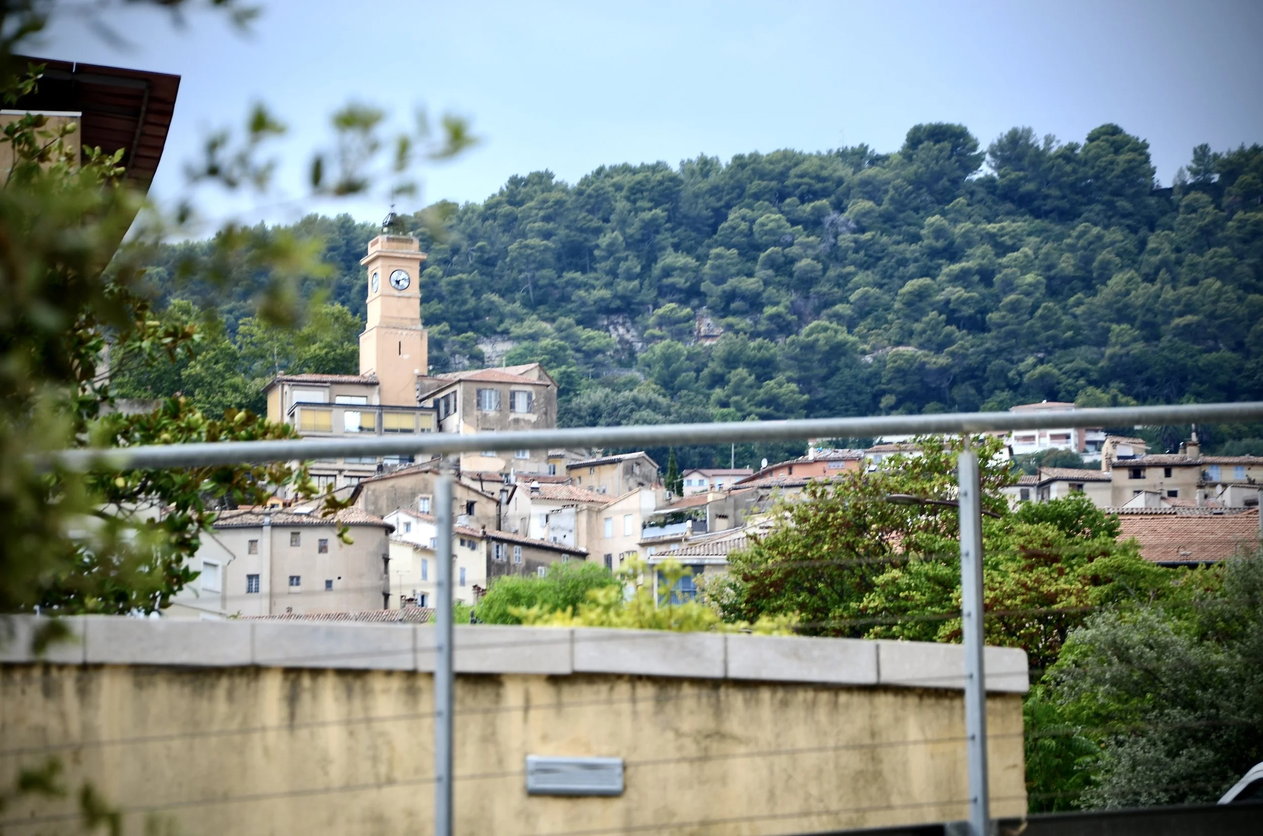 A town with a clock tower in front of a forested hillside, with a balcony railing and partly blurred foliage in the foreground.