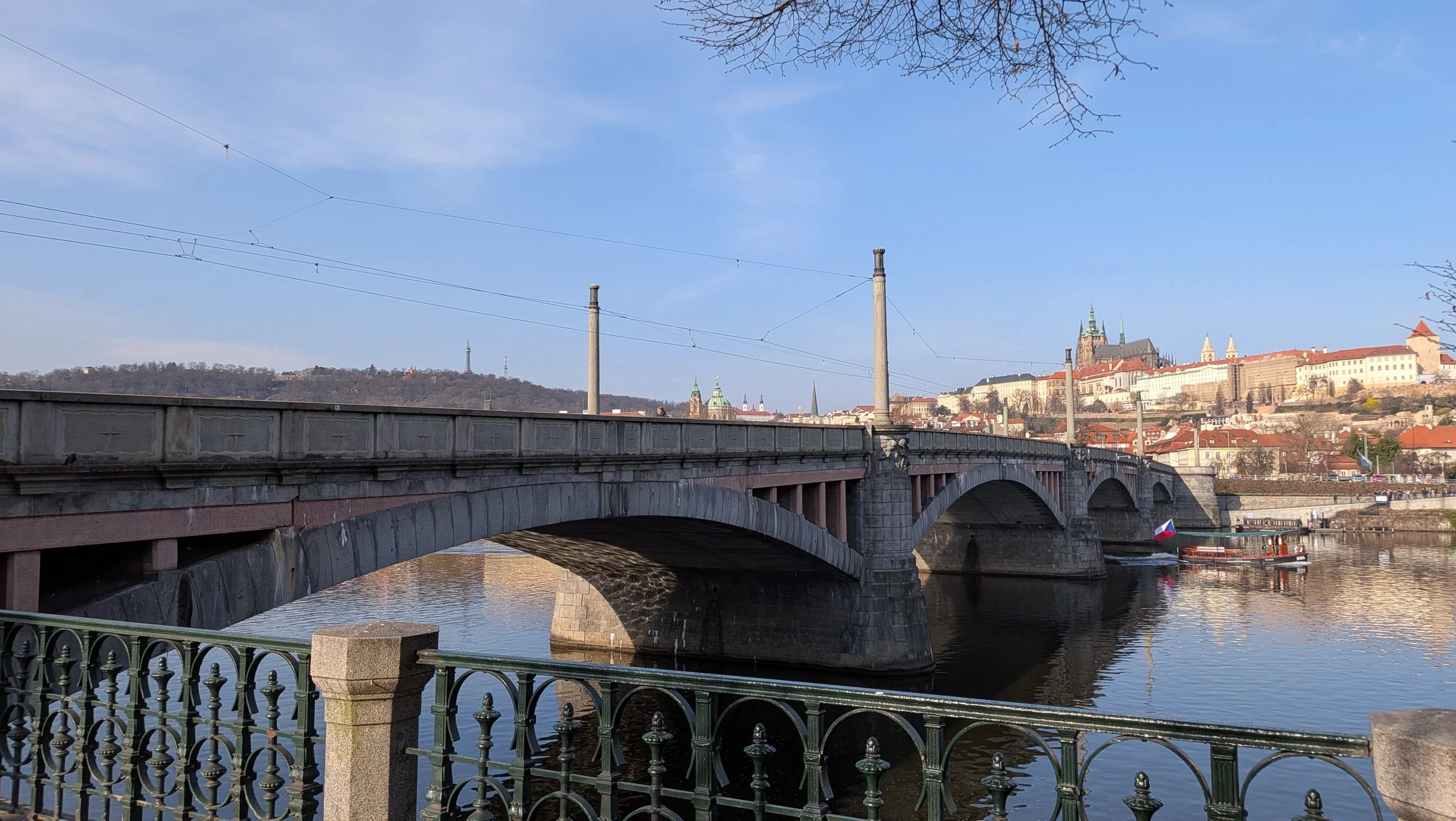 A cityscape view of Prague city with a bridge crossing over a river, historic buildings, and a castle on a hill in the background under a clear blue sky.