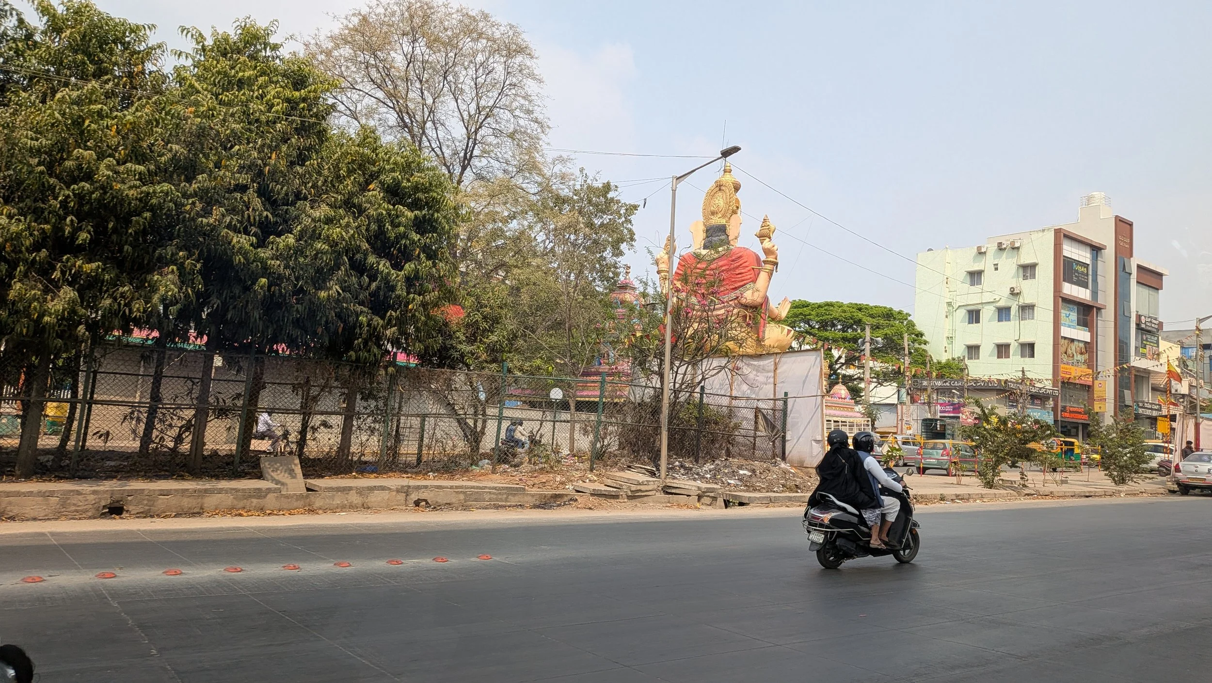 A busy street scene with a large Hindu goddess sculpture in the background, trees, and several multi-story buildings. A person wearing a helmet is riding a scooter on the street.