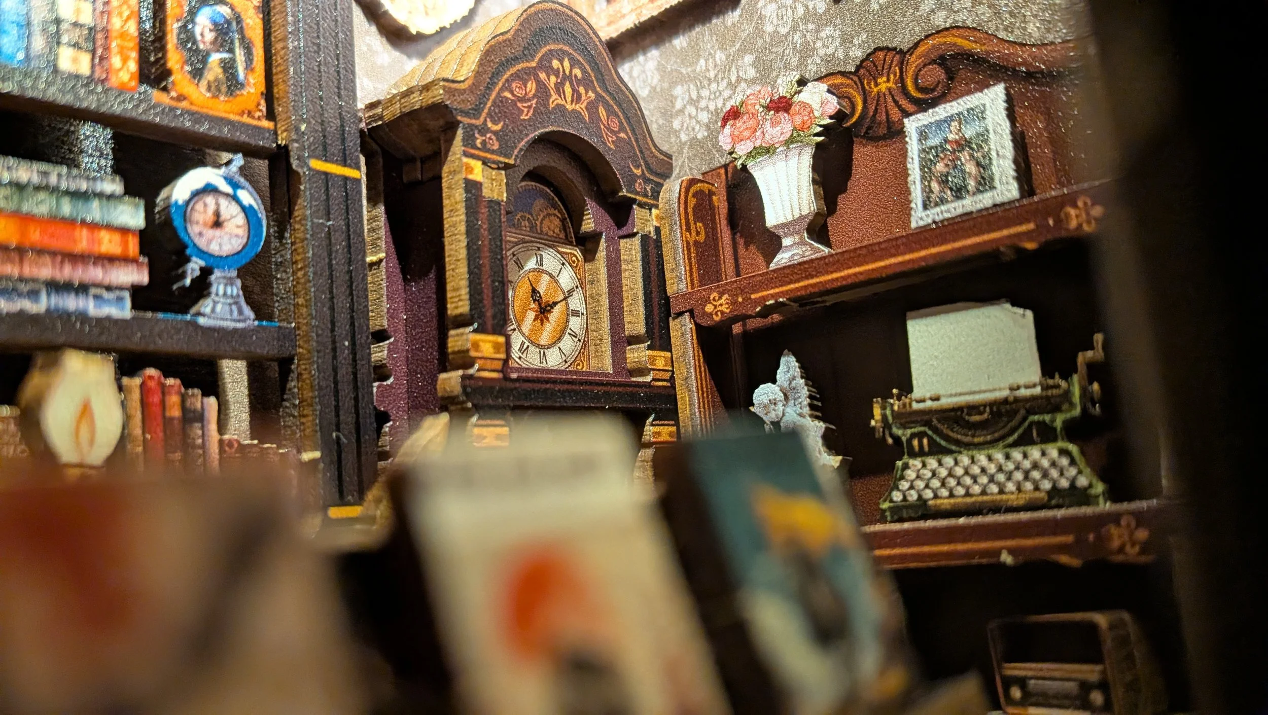 A close-up image showing a miniature bookshelf with a decorative clock, a flower vase, and other small items. The background has a romantic, vintage style.