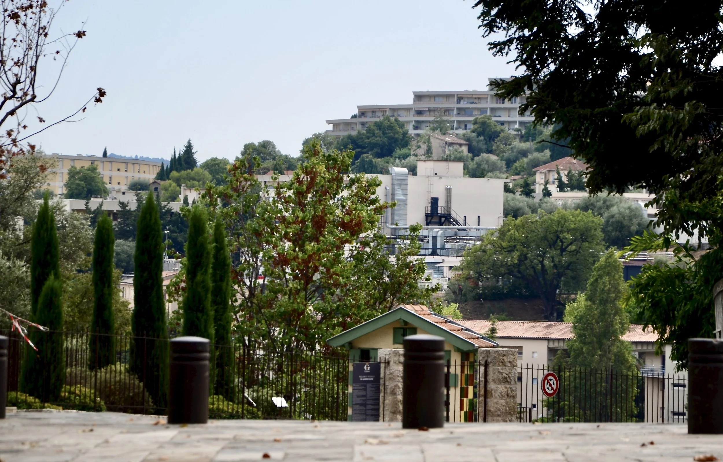 A cityscape with trees, buildings, and a hillside residential area in the background, seen from a paved area with bollards, a fence, and some signage in the foreground.
