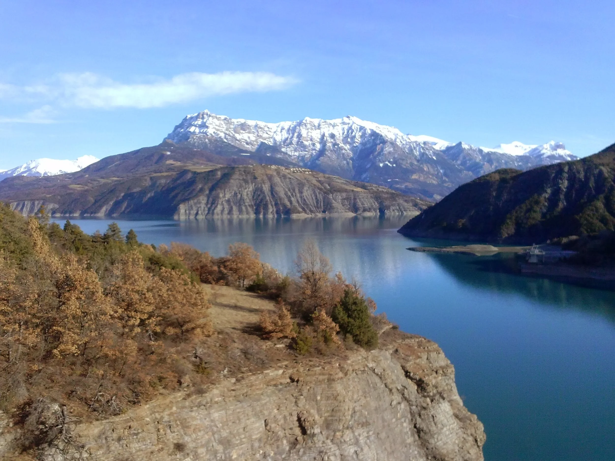 Snow-capped mountains overlooking a calm lake with some trees and rocky shoreline in the foreground.