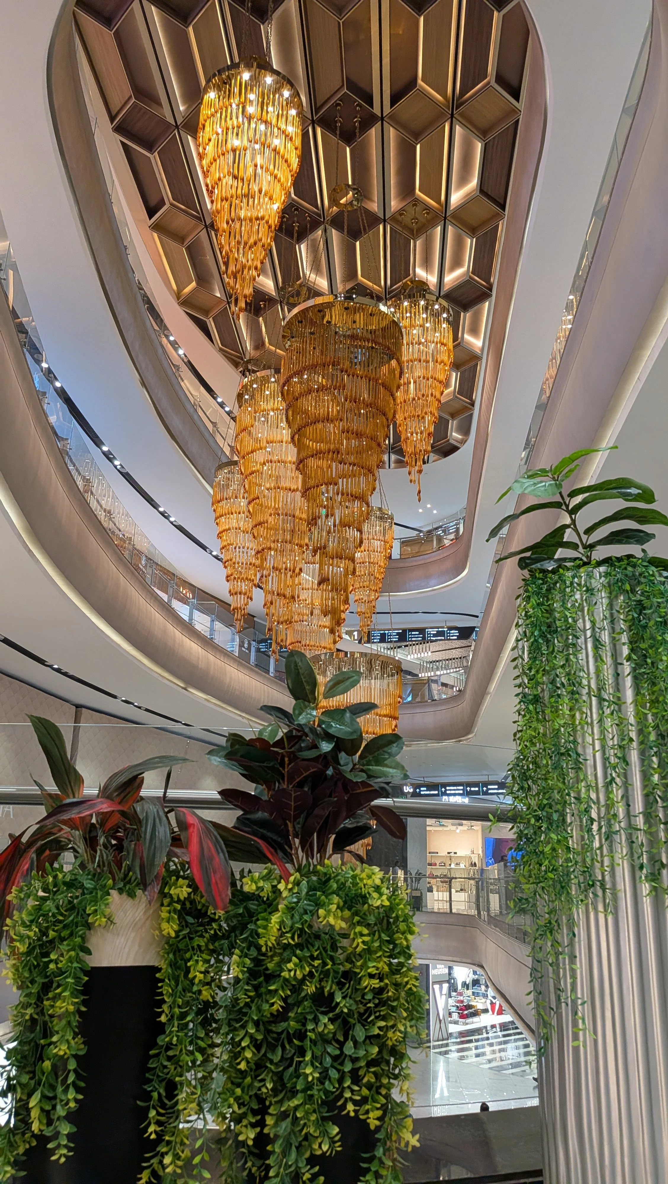 Interior of a shopping mall with multiple golden chandeliers hanging from a ceiling with honeycomb-patterned design, surrounded by green plants in the foreground and shops in the background.