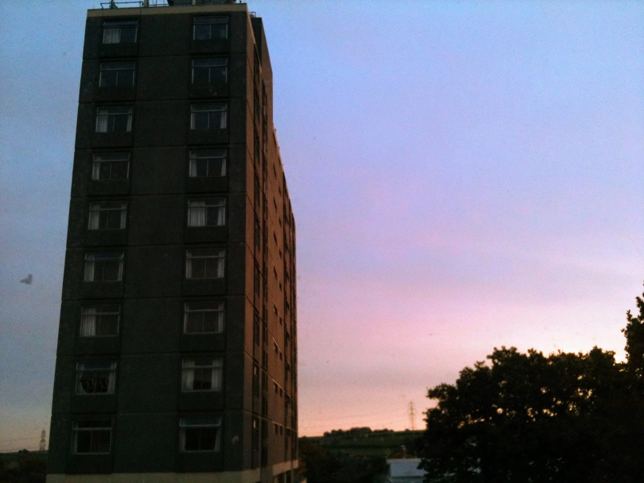 A tall residential apartment building during twilight with dark exterior walls and multiple windows. The sky is transitioning from dusk to night with soft pastel colors, and there are trees silhouetted at the bottom right corner.
