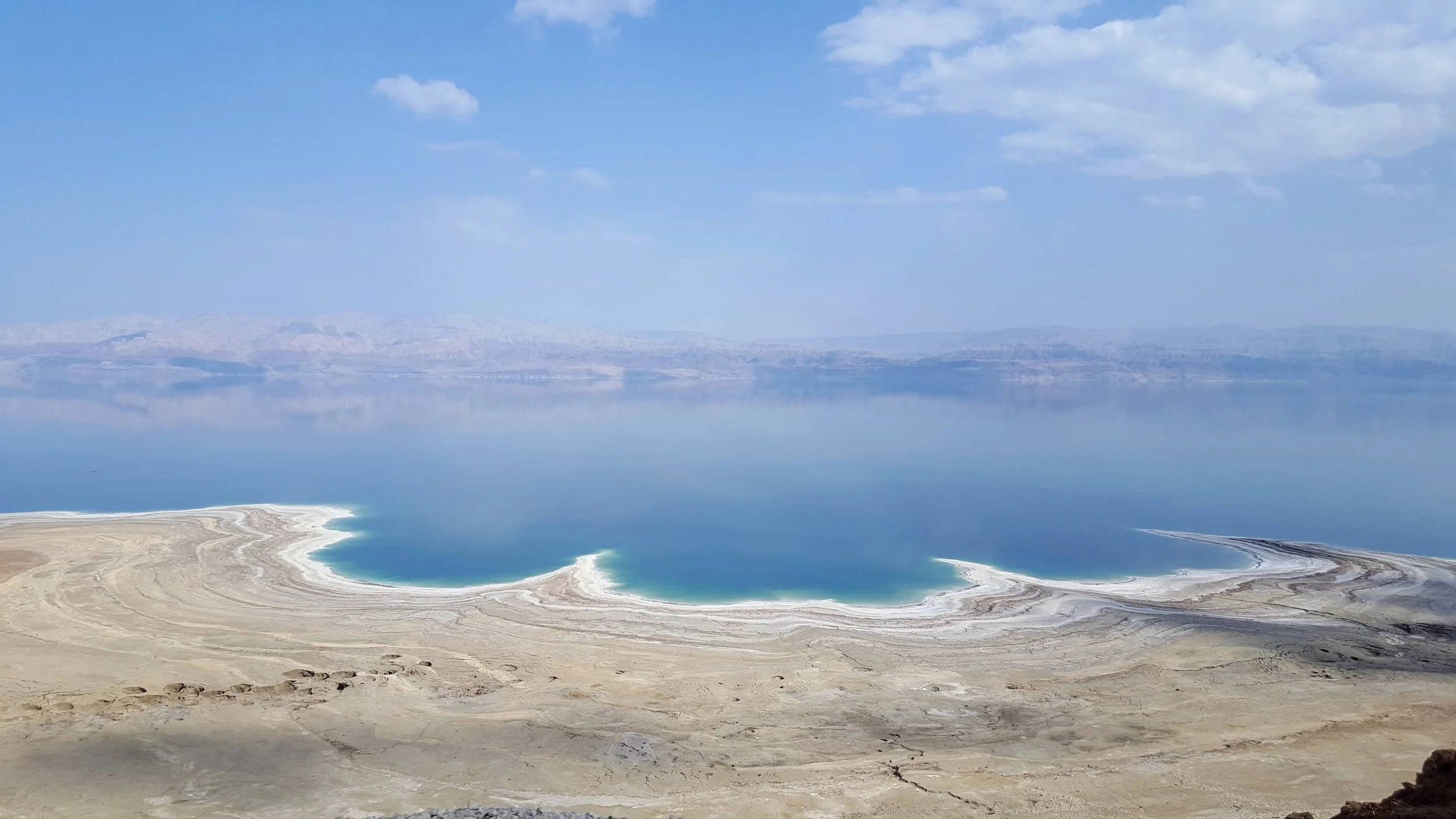 Salt flats and a blue lake with mountains in the distance under a partly cloudy sky.