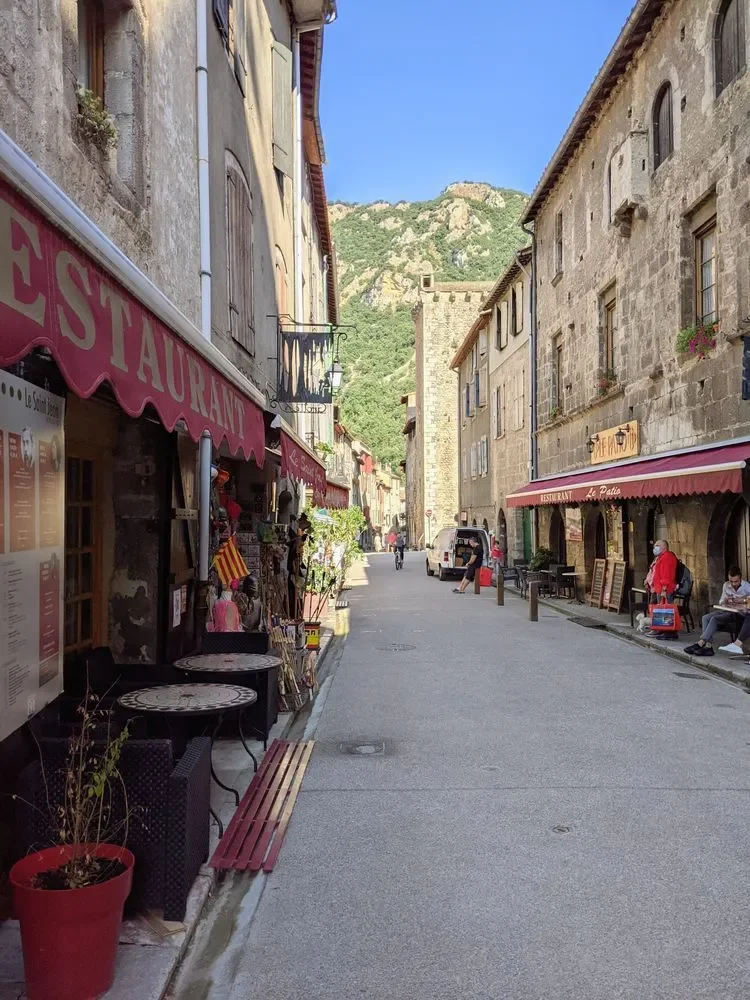 A narrow street in a European town with stone buildings, outdoor seating, and a mountain in the background.