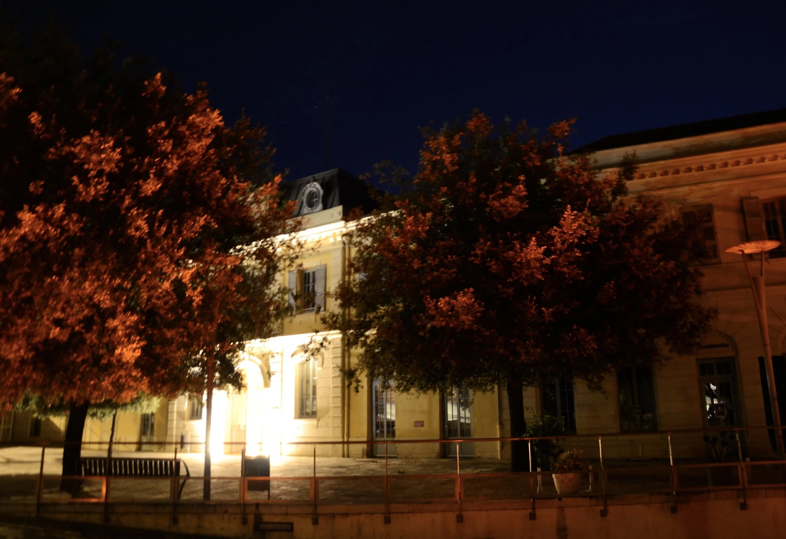 Nighttime scene of a historic building with illuminated windows and a clock tower, partially obscured by trees with orange and red autumn leaves.