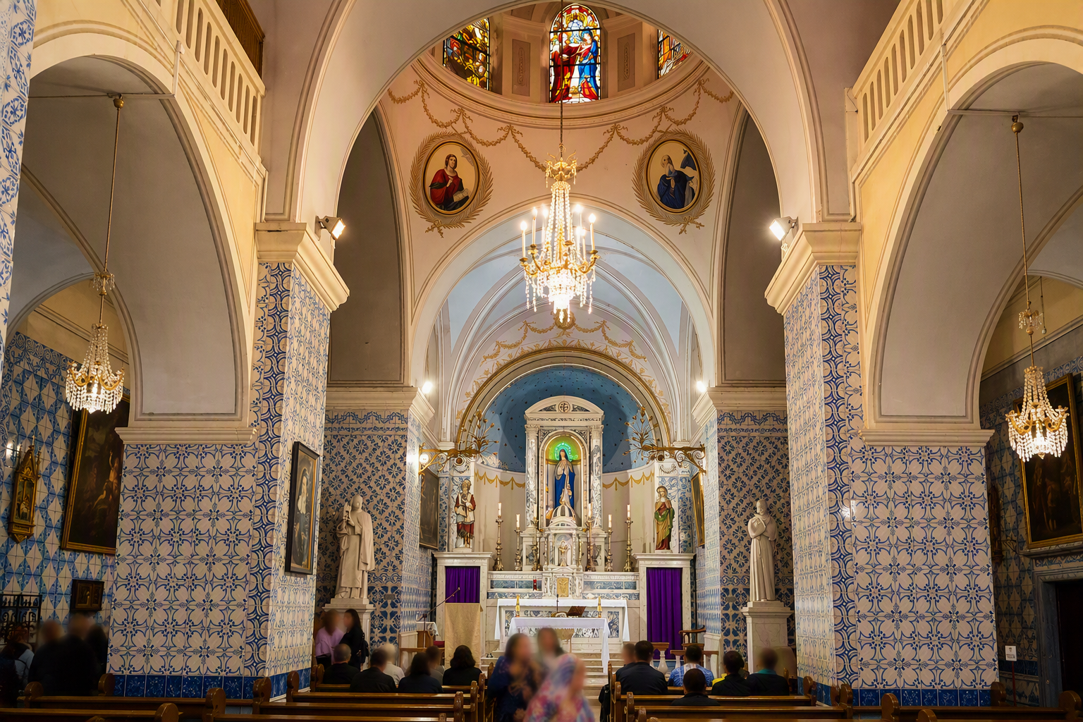 Interior of a church with an ornate altar, religious statues, stained glass windows, and chandeliers, with congregation seated in pews.