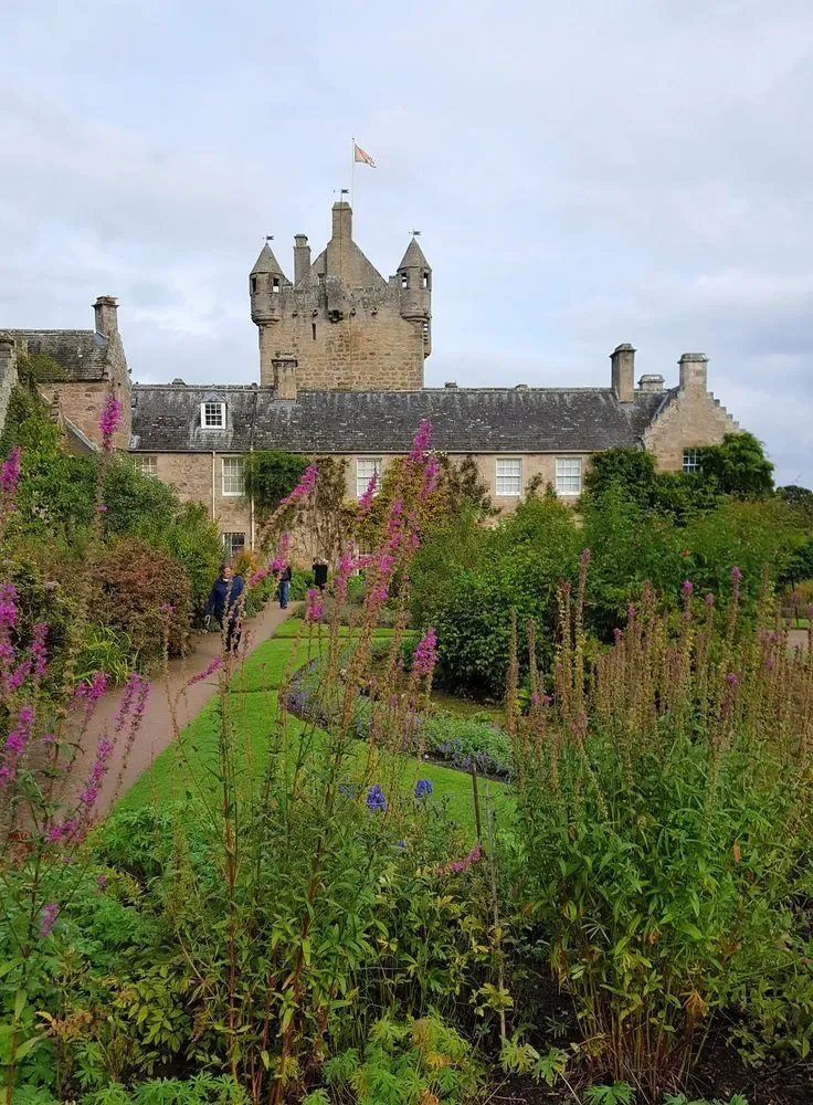 A historic stone castle with towers and a flag on top, surrounded by a lush garden with flowering plants and a walking path, under a cloudy sky.
