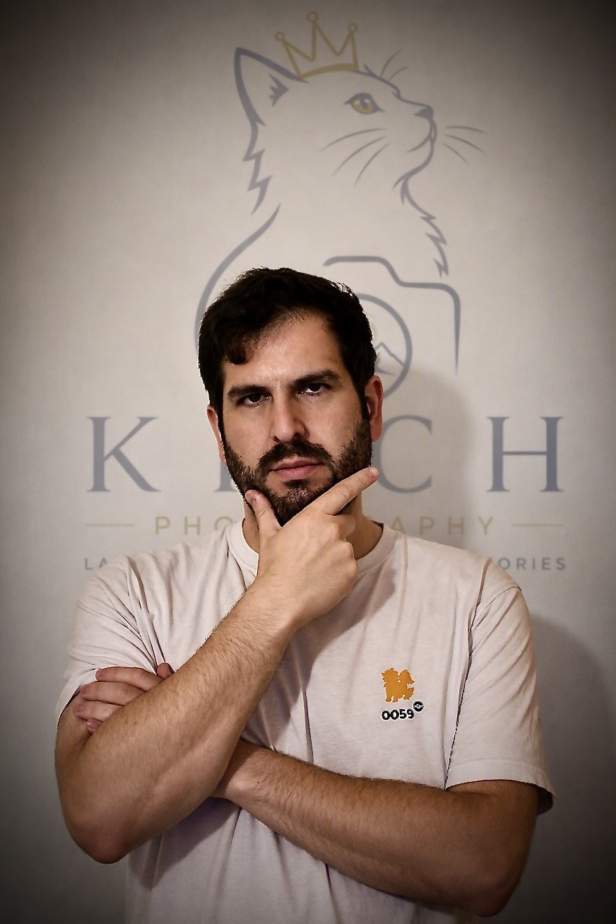 A man with dark hair and beard standing in front of a wall with a logo of a cat wearing a crown and the words 'KITCH' and 'PHOTOGRAPHY'. The man is wearing a light-colored t-shirt with an orange lion graphic and the number 0059. He is posing with one hand on his chin and the other arm crossed.