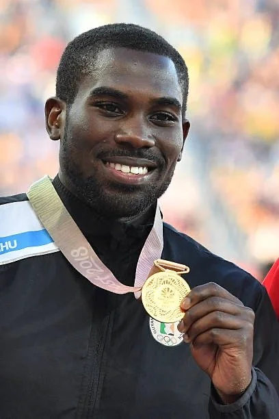 Man smiling and holding up a gold medal at an outdoor event.