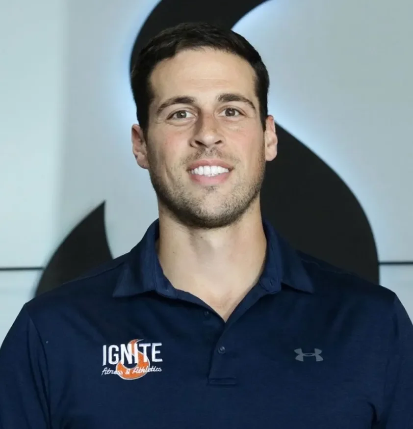 A young man with short dark hair and light skin, smiling, wearing a navy blue collared shirt with 'IGNITE Fitness & Athletics' logo and Under Armour logo, standing in front of a light-colored background with a large black and white logo behind him.