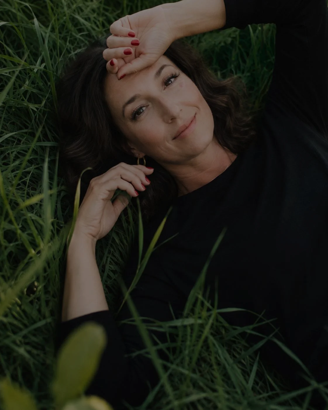 A woman with dark brown hair and red nail polish lying in green grass, smiling softly at the camera.