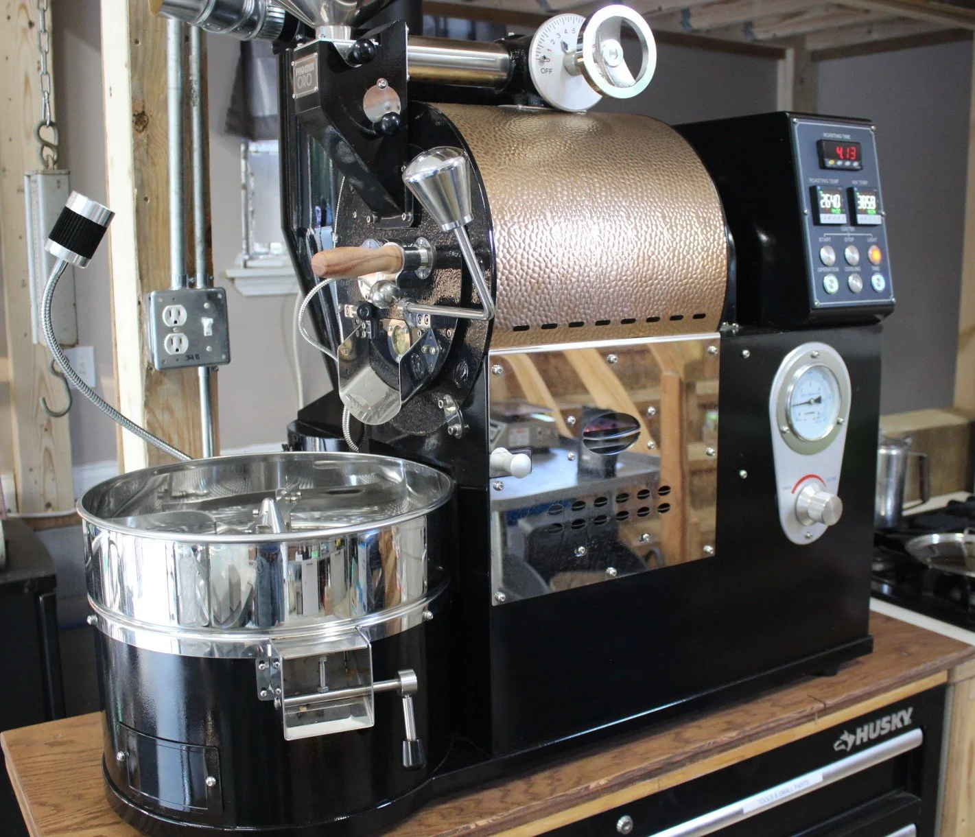 A commercial roaster machine used for roasting coffee beans, with a metal drum, control dials, and a temperature gauge, placed on a wooden table in a workshop.