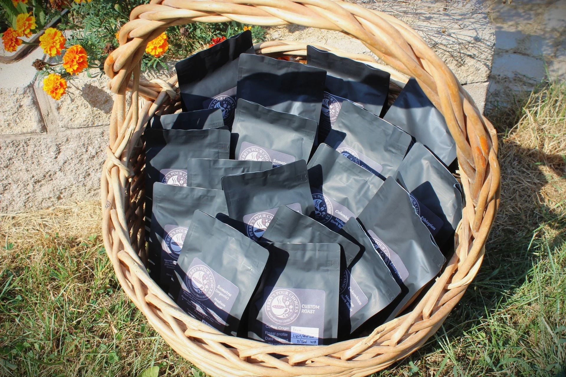 A woven basket outdoors filled with black coffee bags on grassy ground near a brick wall with orange marigold flowers nearby.