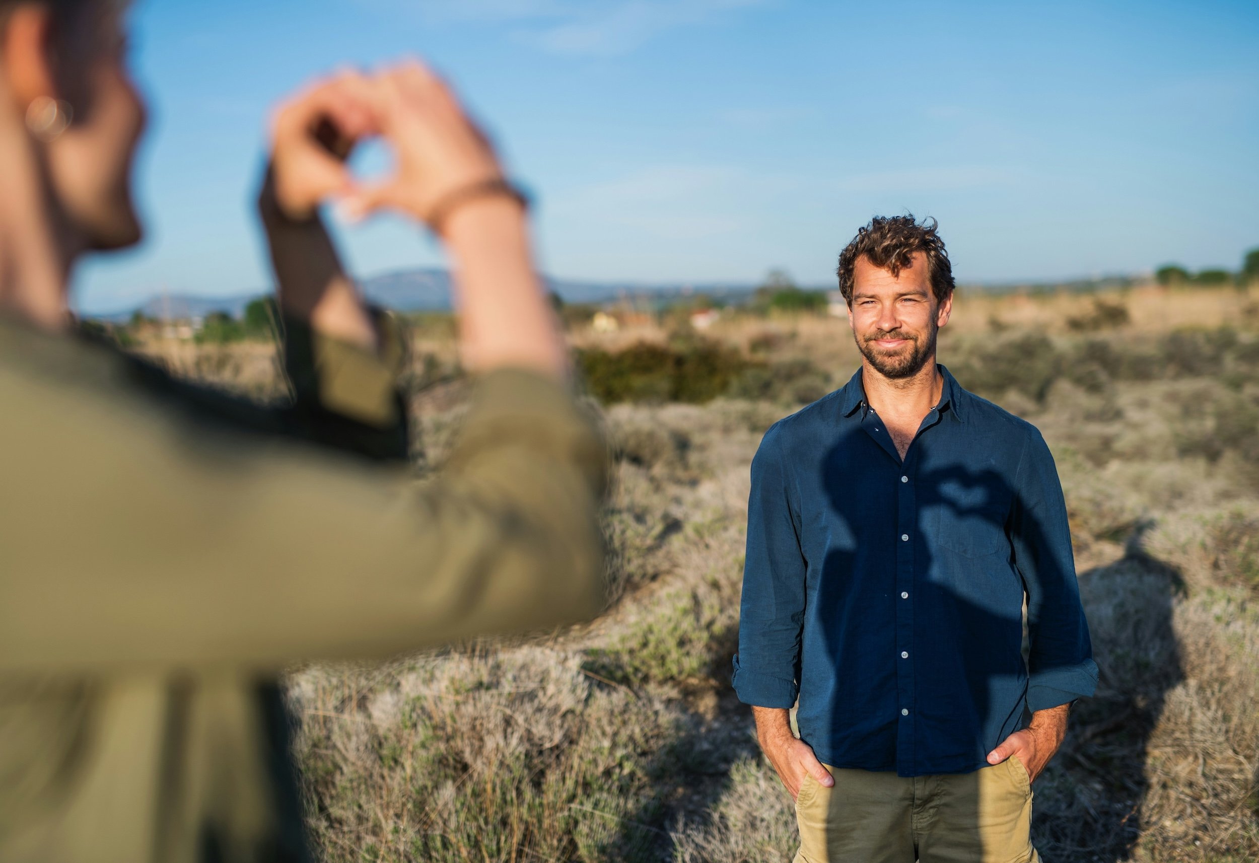 A man in a blue shirt standing outdoors in a field, smiling at the camera. Another person is taking the photo, with their hands visible in the foreground forming a heart shape.
