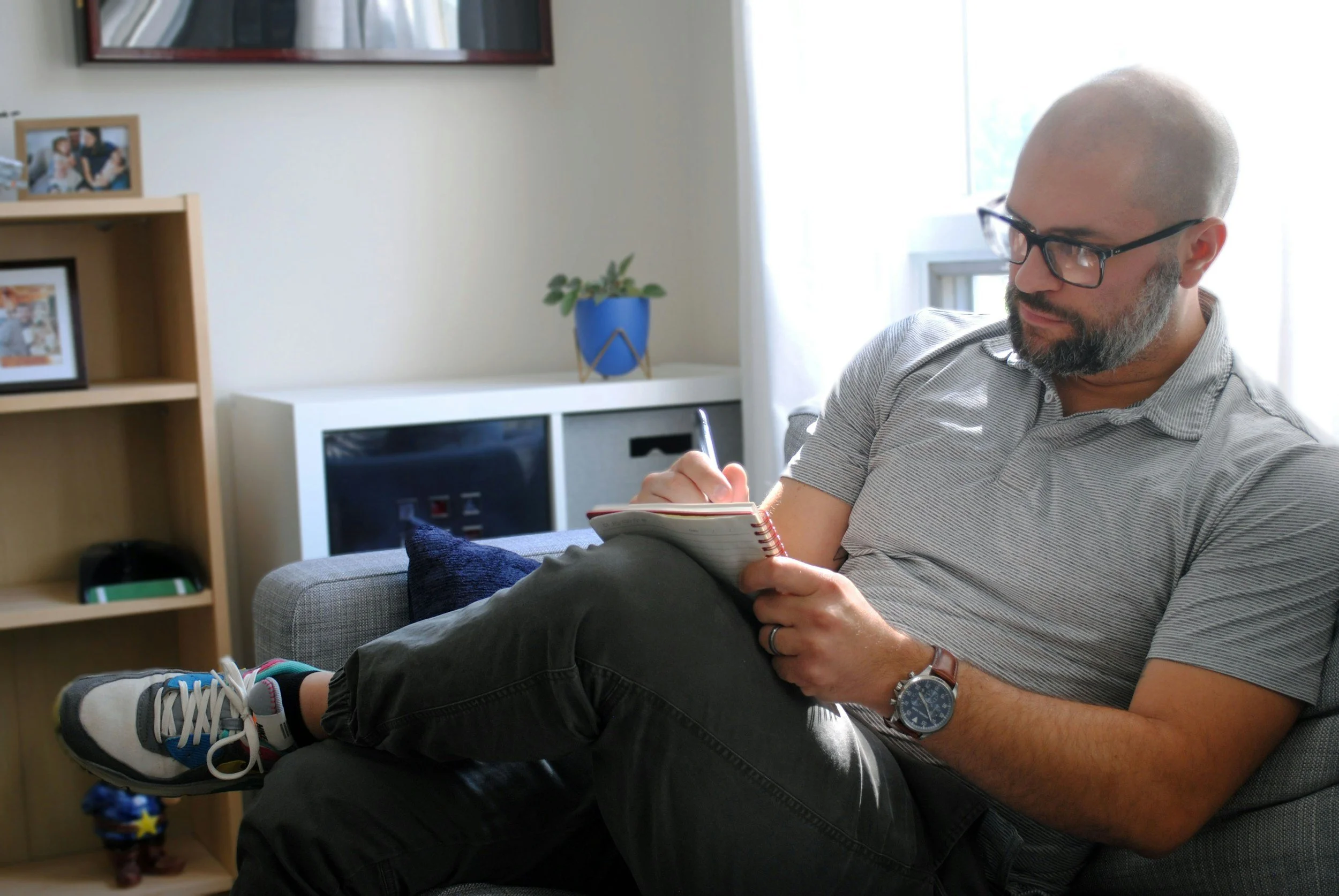 A bald man with glasses, a beard, and a wristwatch is sitting on a gray sofa, drinking from a mug while writing in a notebook. The room has a window with white curtains, a wooden bookshelf with family pictures, and a blue potted plant.