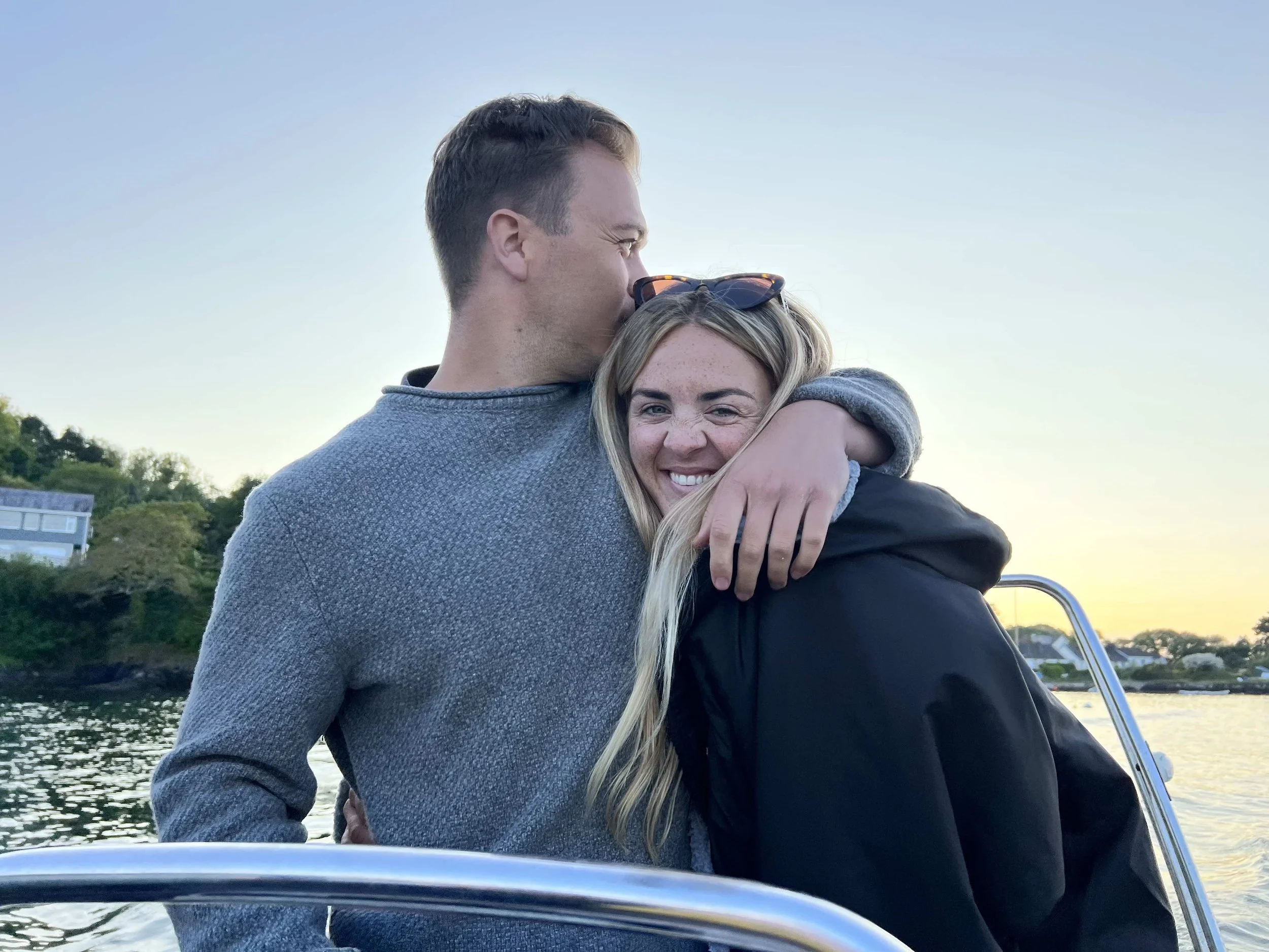 A man and woman sharing a hug on a boat during sunset, with trees and modern buildings in the background.