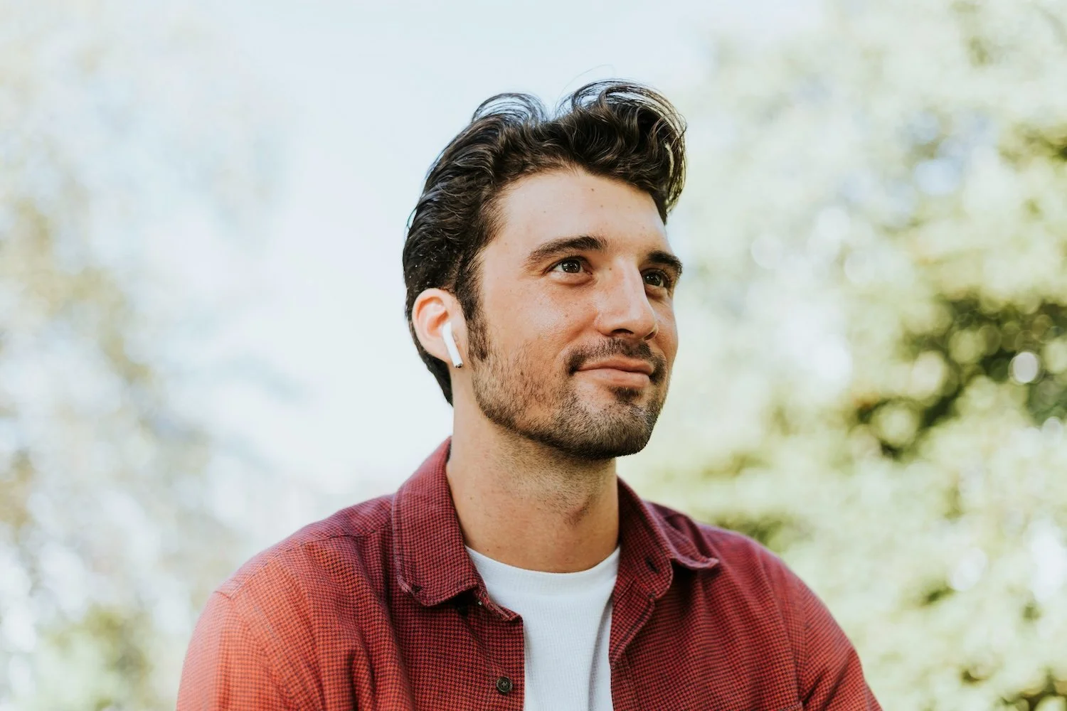 A man with dark hair and facial hair, wearing a white shirt and a red plaid shirt, is outdoors with trees in the background. He has a calm expression and is wearing wireless earbuds.