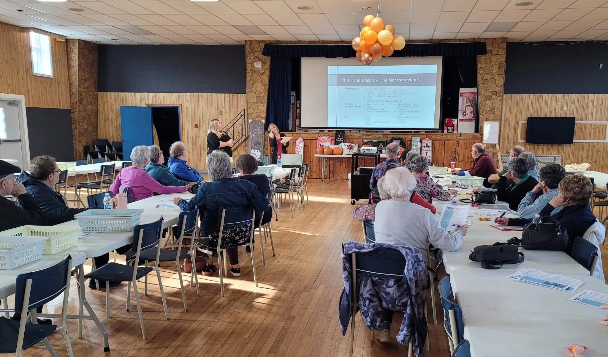 Seniors at long tables listening to a presentation in a community hall decorated with balloons