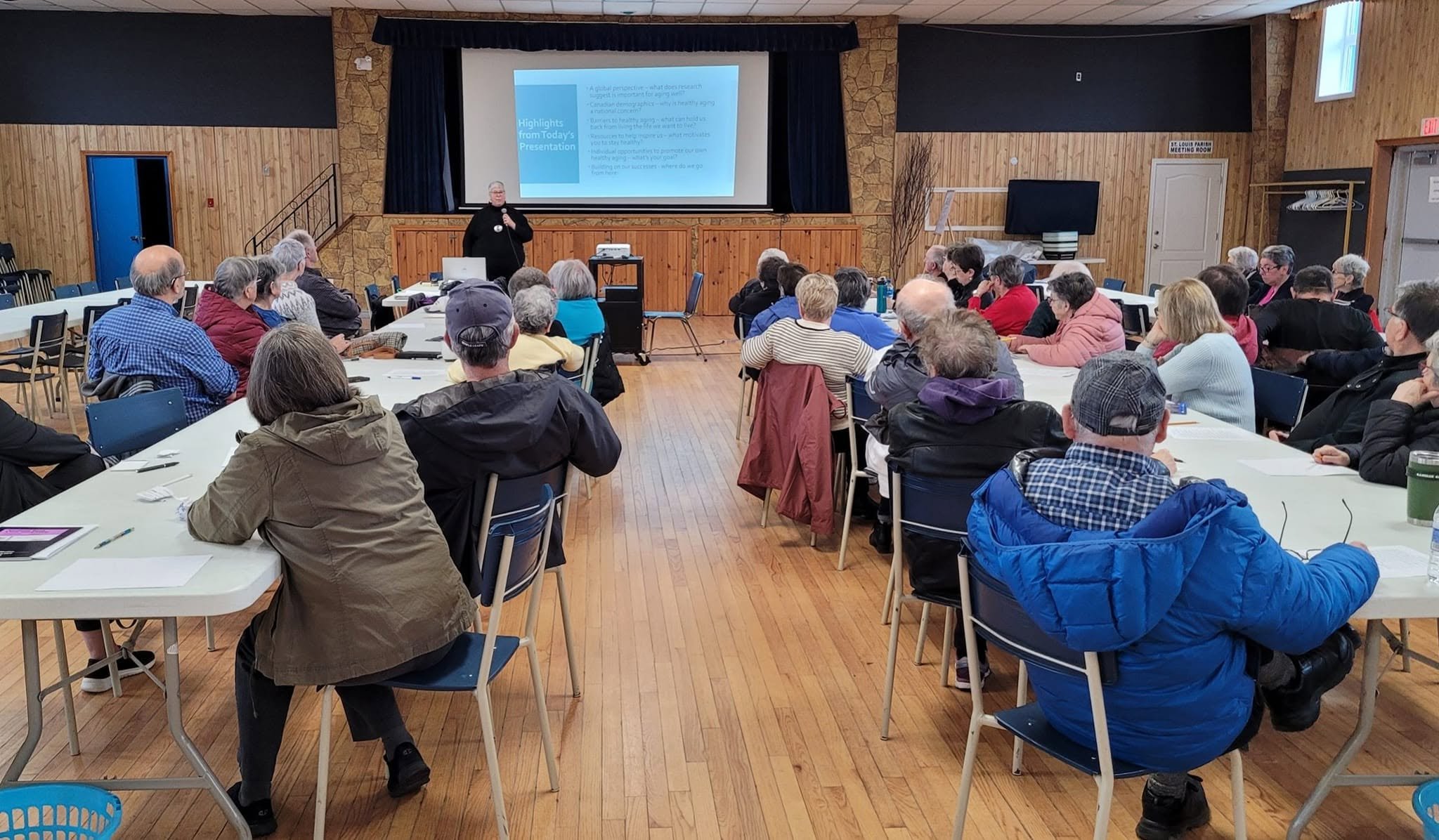Older adults seated at tables in a community hall watching a presentation at a STAC meeting