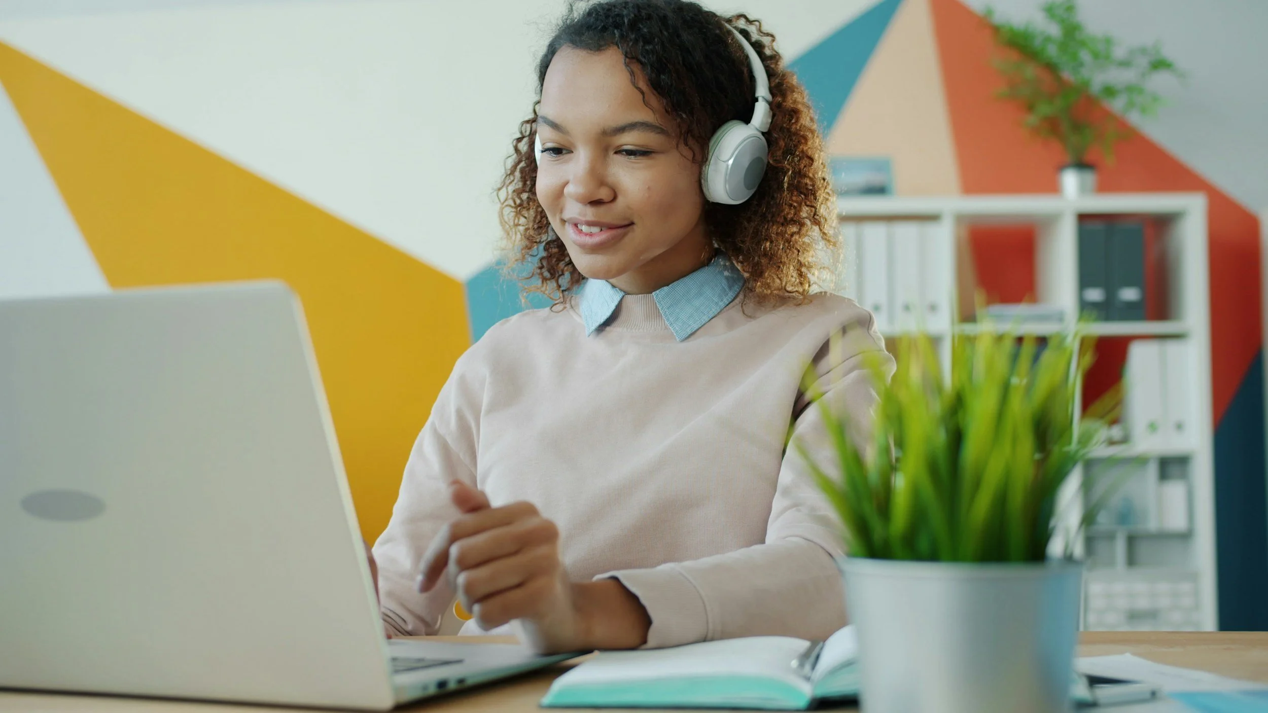 A woman wearing headphones is working on a laptop in a colorful, modern office space with a bookshelf, a potted plant, and geometric wall decor.