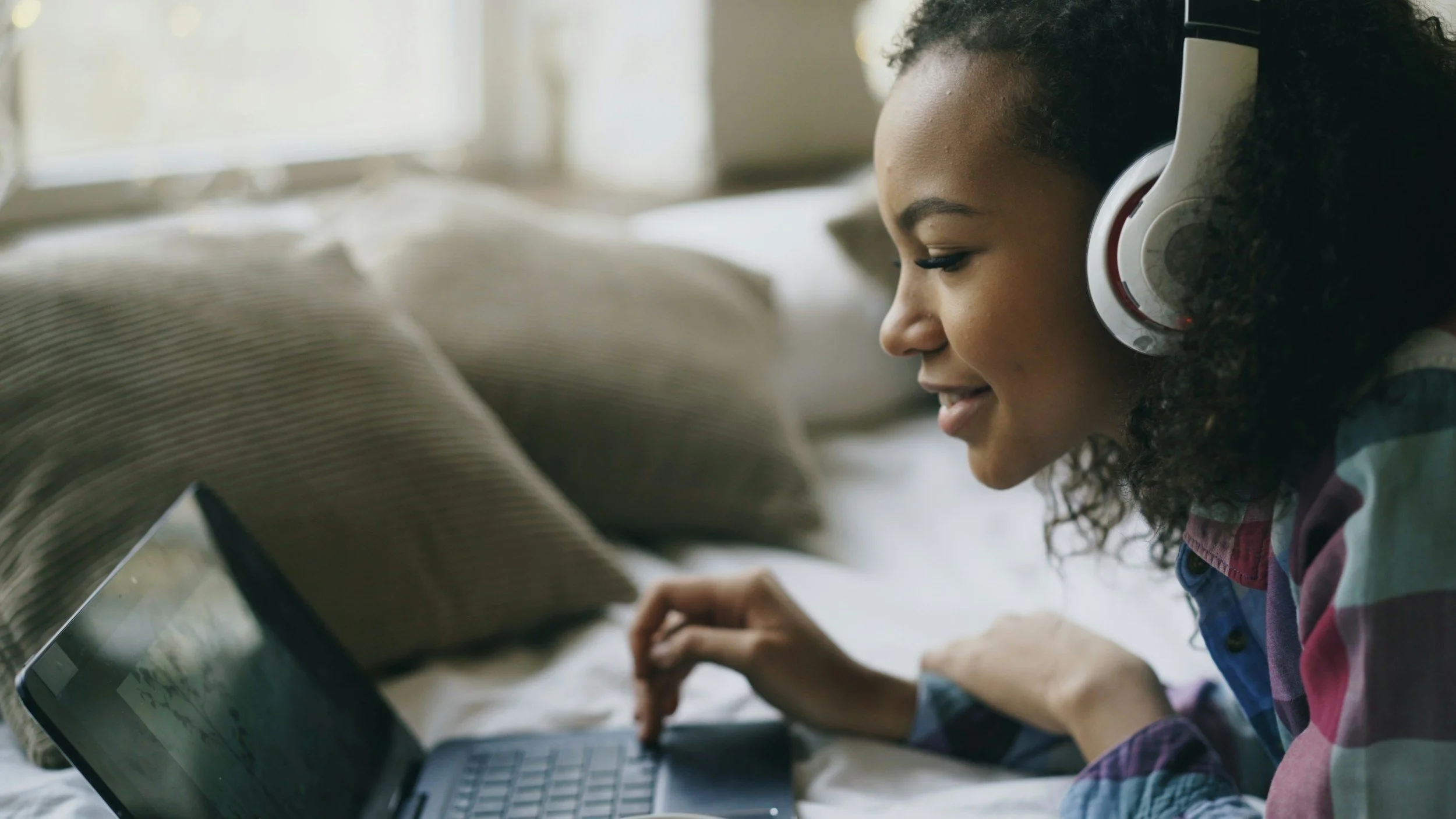 Young girl with curly hair wearing headphones, smiling, using a laptop.