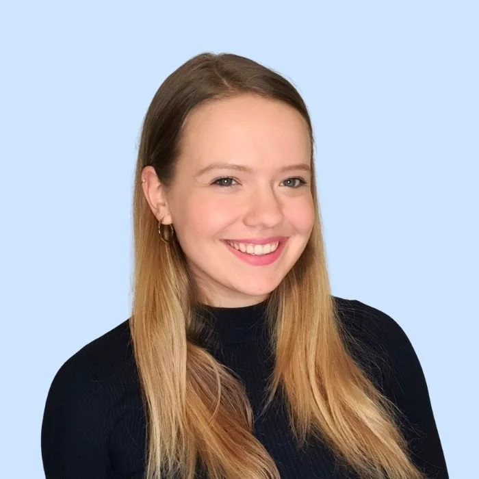 A young woman with long blonde hair, smiling, wearing a black top, against a light blue background.