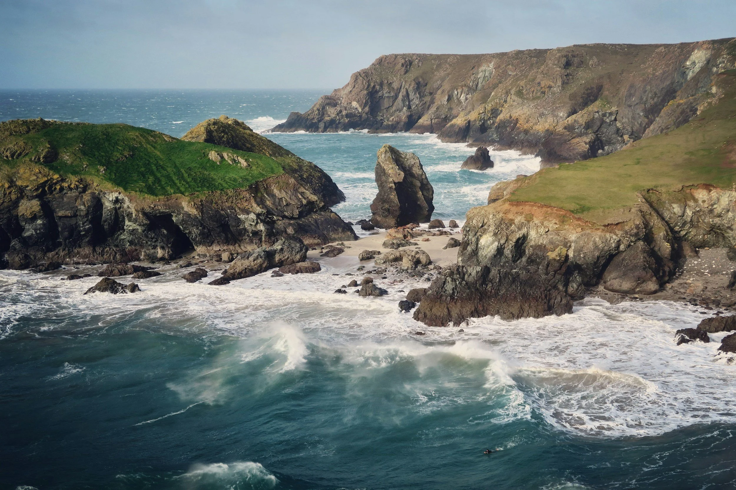 Rocky coastline with green patches, ocean waves crashing against rocks, and cliffs in the background under a cloudy sky.