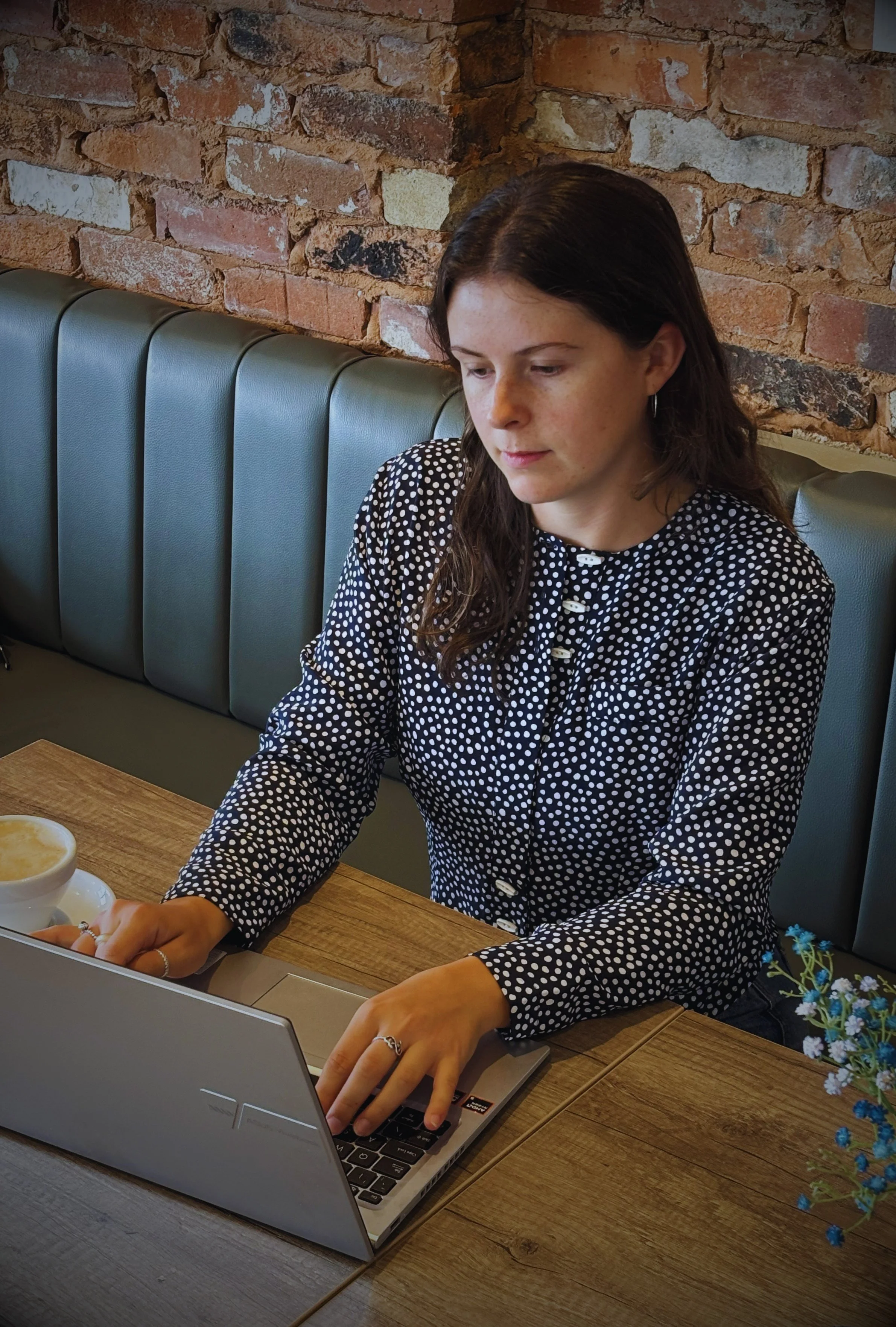 Bookkeeper in a polka-dot blouse working on a laptop at a wooden café table against a brick wall