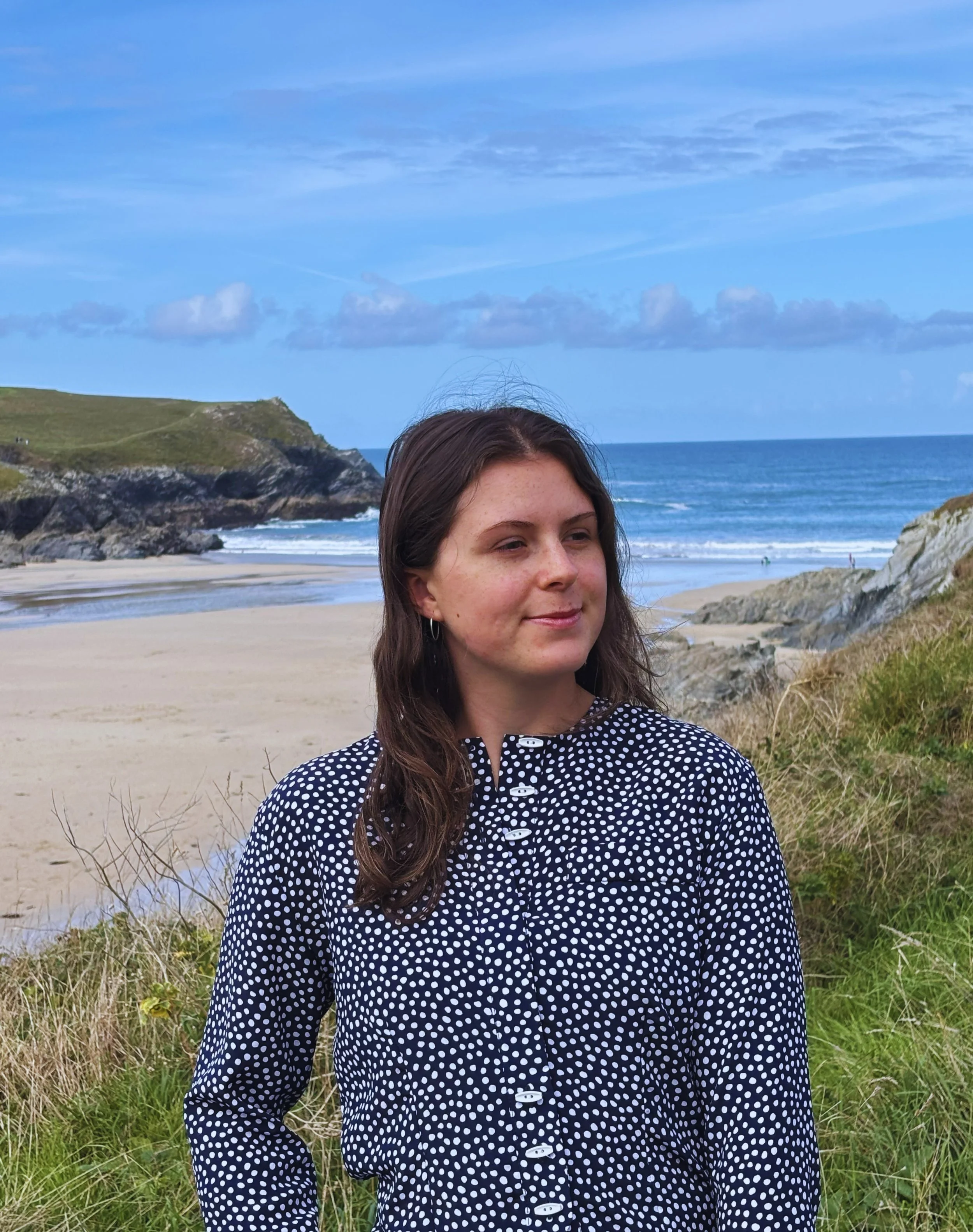 Bookkeeper in a navy polka-dot blouse posing at a sandy beach with rocky cliffs and ocean waves in Cornwall