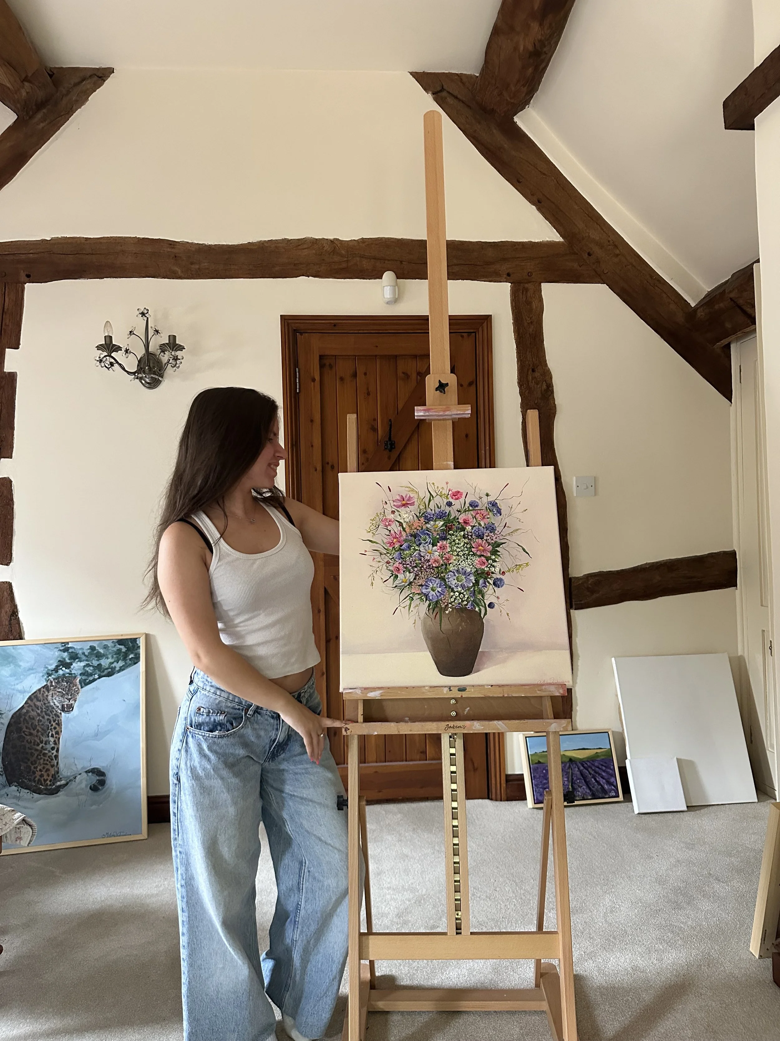 A woman in a white tank top and blue jeans standing in a room with wooden beams on the ceiling, holding a large canvas of a colorful flower bouquet painting on an easel.