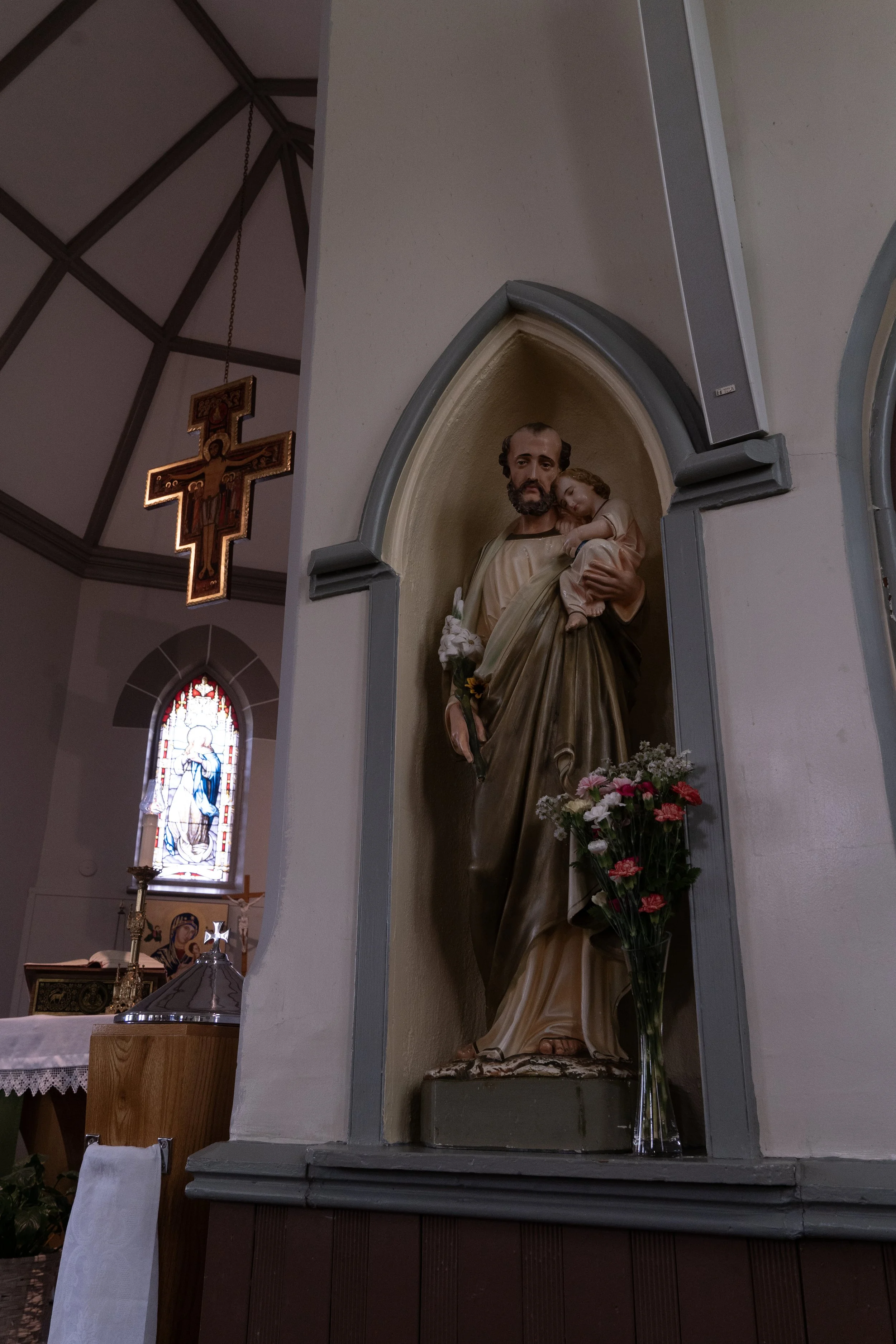 Religious altar with a statue of St. Joseph holding baby Jesus, accompanied by a vase of flowers, and a carved crucifix hanging in the church interior. Stained glass windows are visible in the background.