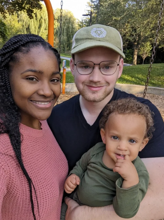 Happy family of three outdoors in a park on a swing set, with trees and grass in the background. A woman with braided hair in a pink sweater, a man with glasses and a cap, and a young child with curly hair wearing a green shirt.