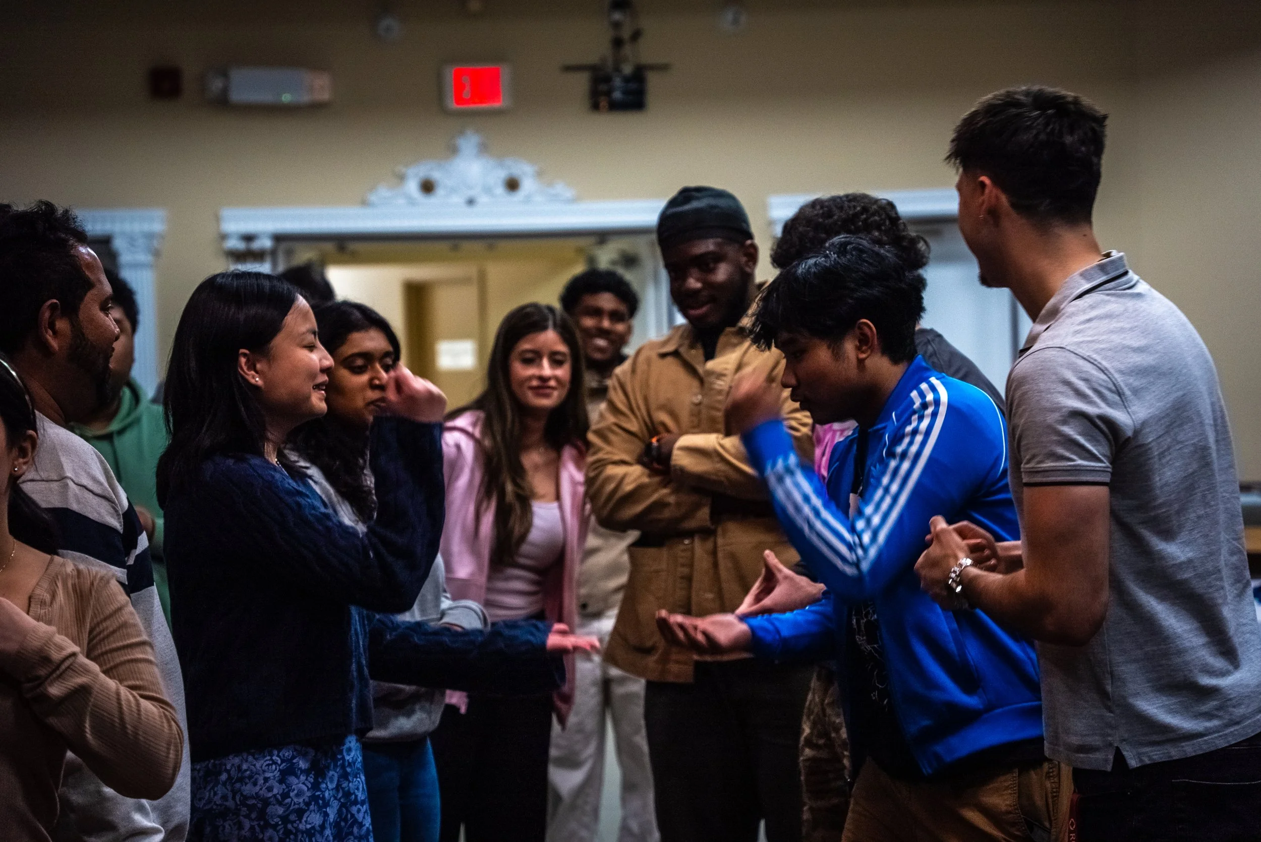 Group of people standing in a semi-circle, engaging in conversation or activity, with a beige wall and door in the background.