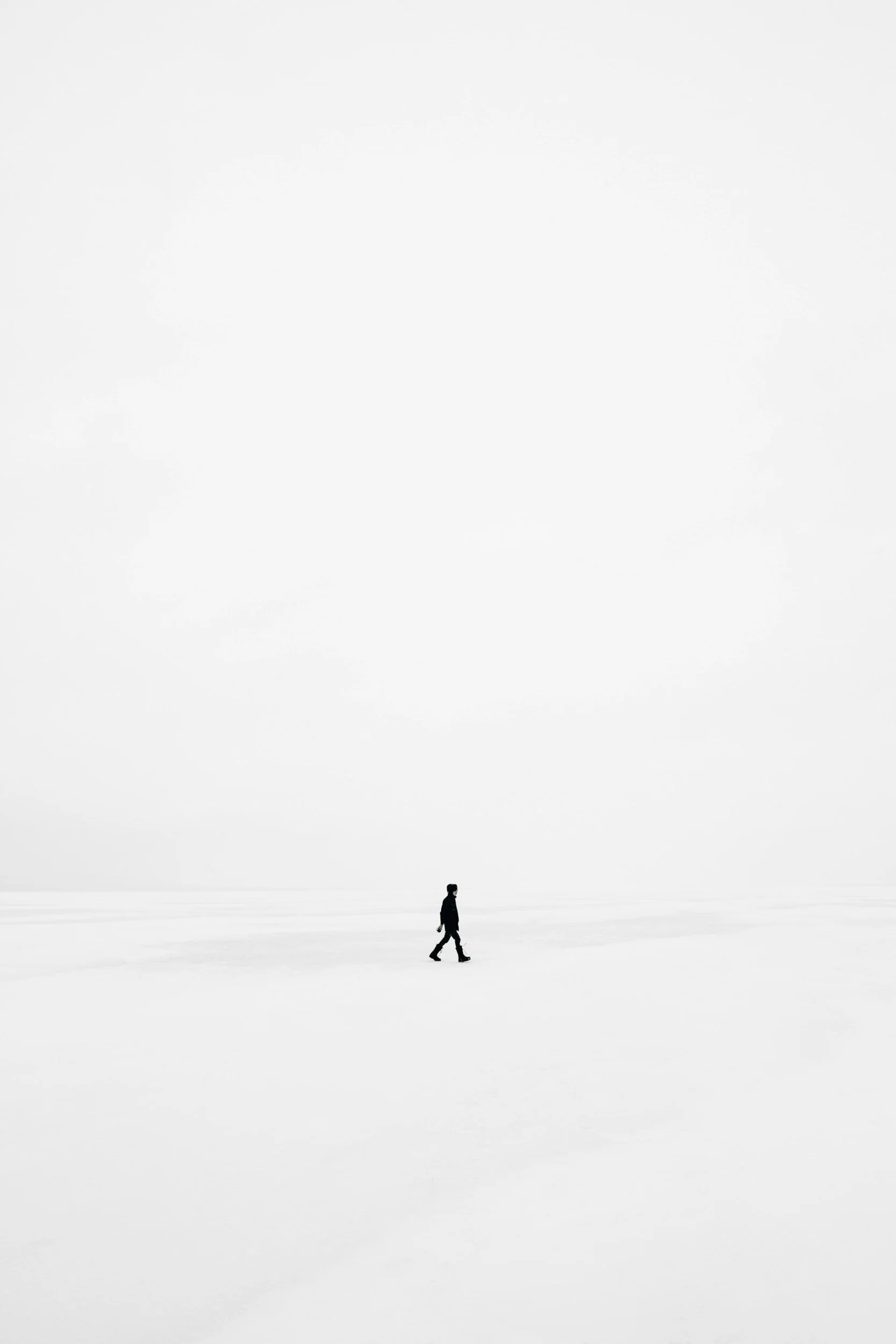A person walking alone on a vast, empty snow-covered landscape with a large, empty, cloudy sky overhead.