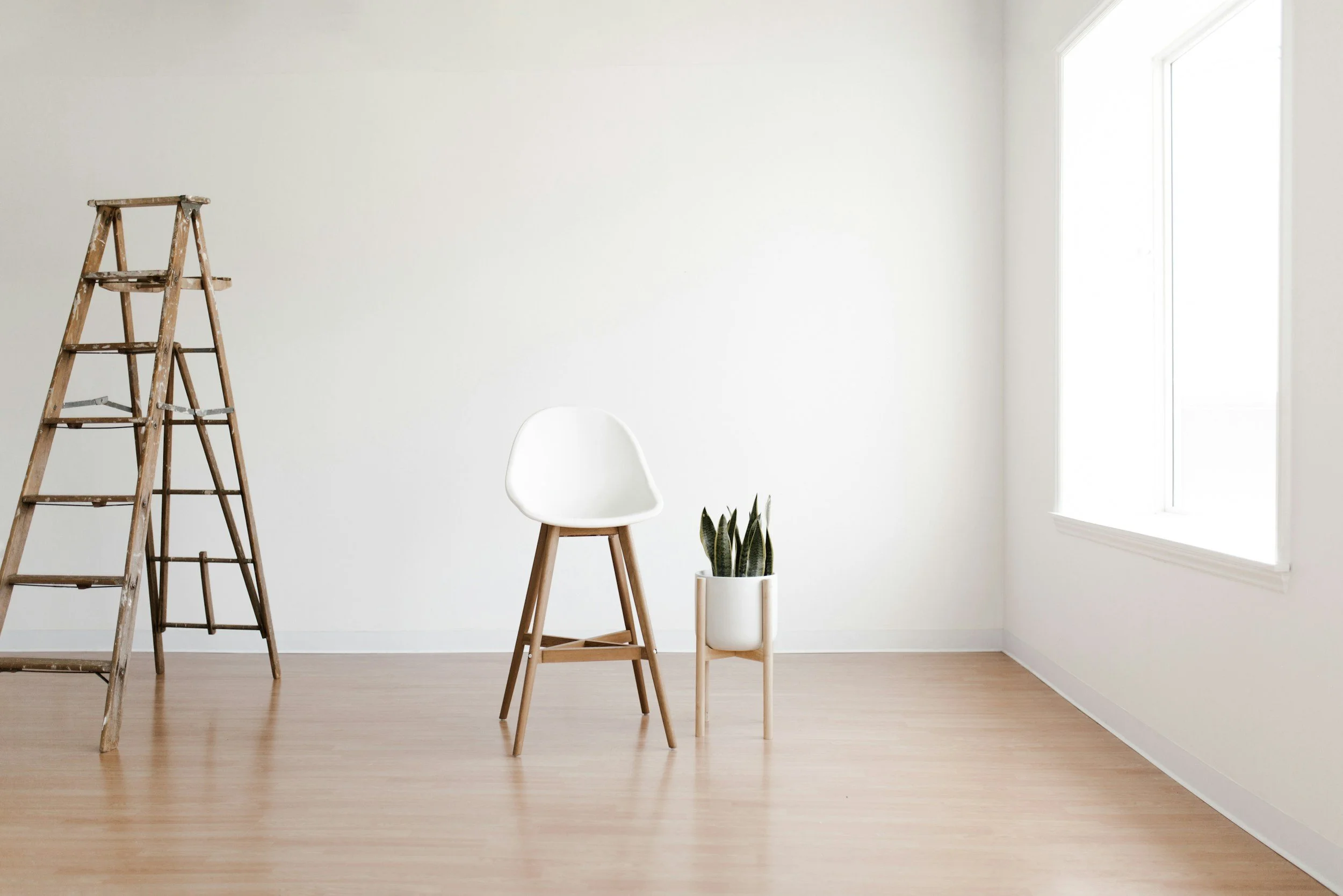 Empty room with wooden flooring, a white wall, a window letting in natural light, a leaning wooden ladder with paint splatters, a white chair, and a potted snake plant.