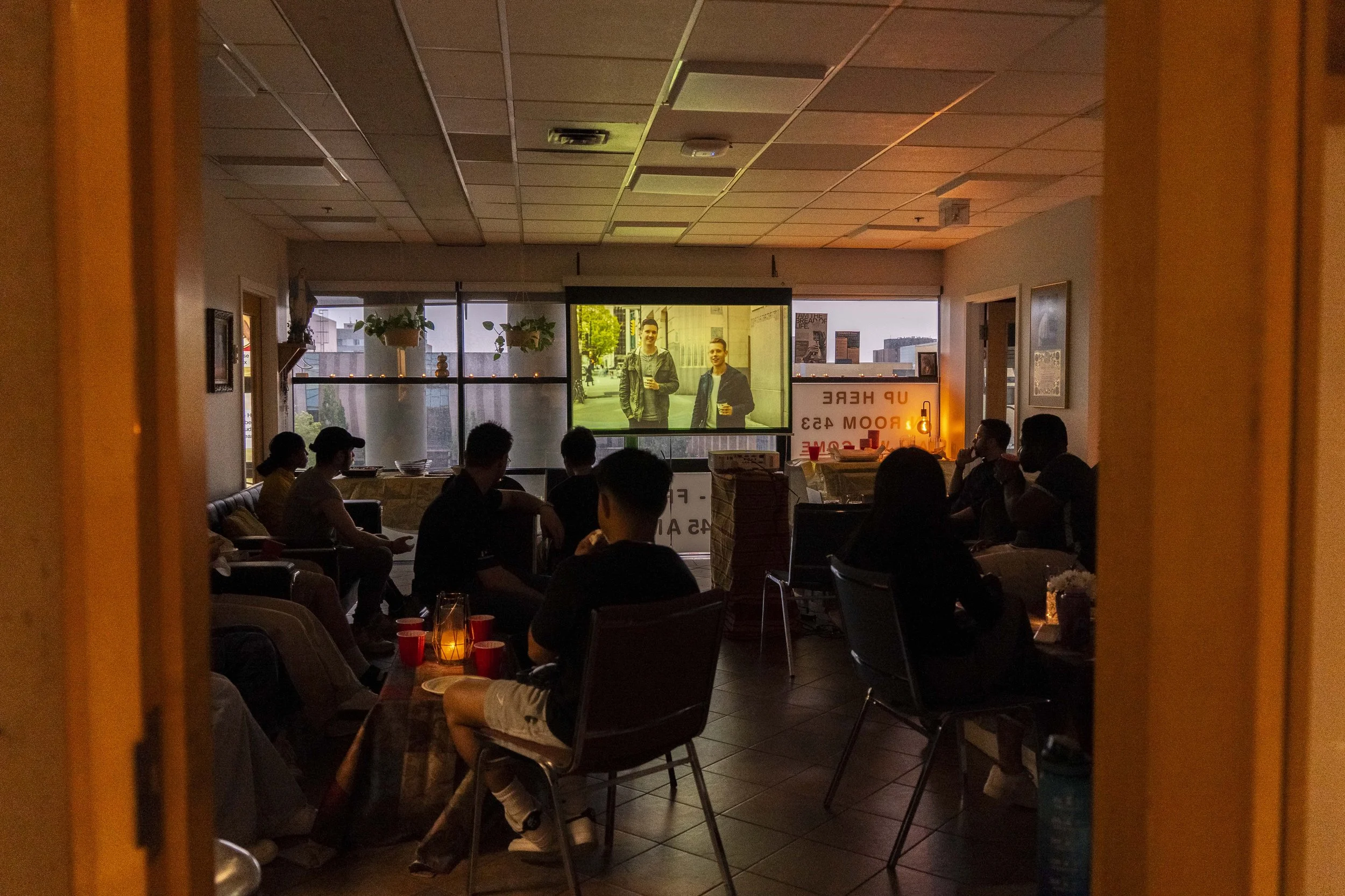 A group of people seated and watching a large screen displaying two men with drinks outside in an urban setting, inside a dimly lit room with windows, plants, and hanging decorations.