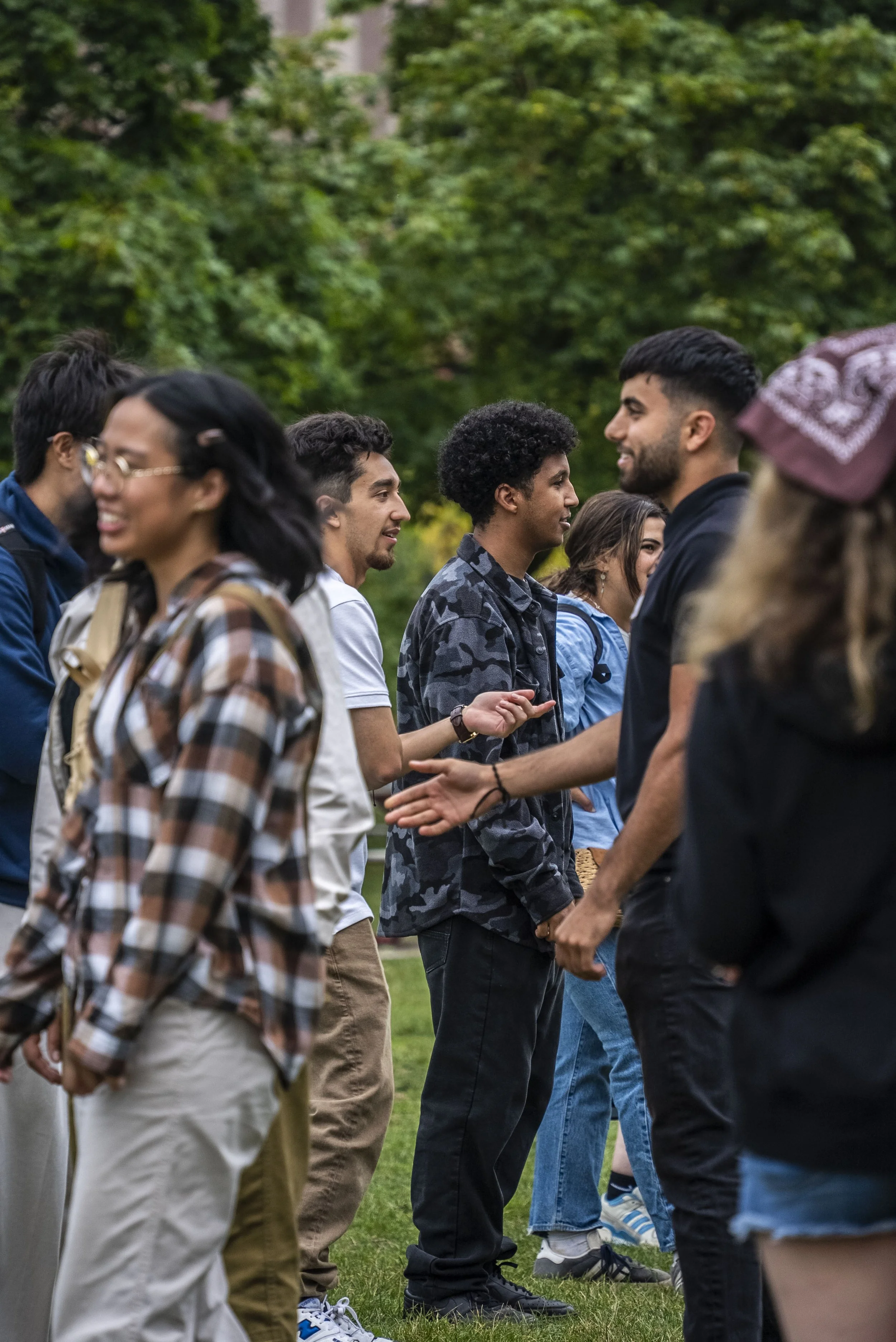 Group of diverse young adults standing in a line outdoors, shaking hands and chatting, with green trees in the background.