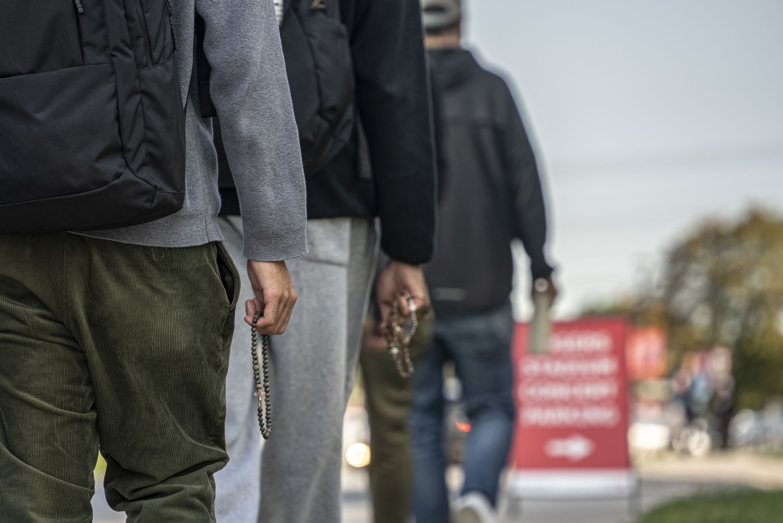 People standing in a line outdoors, holding rosaries, some with backpacks and casual clothing.