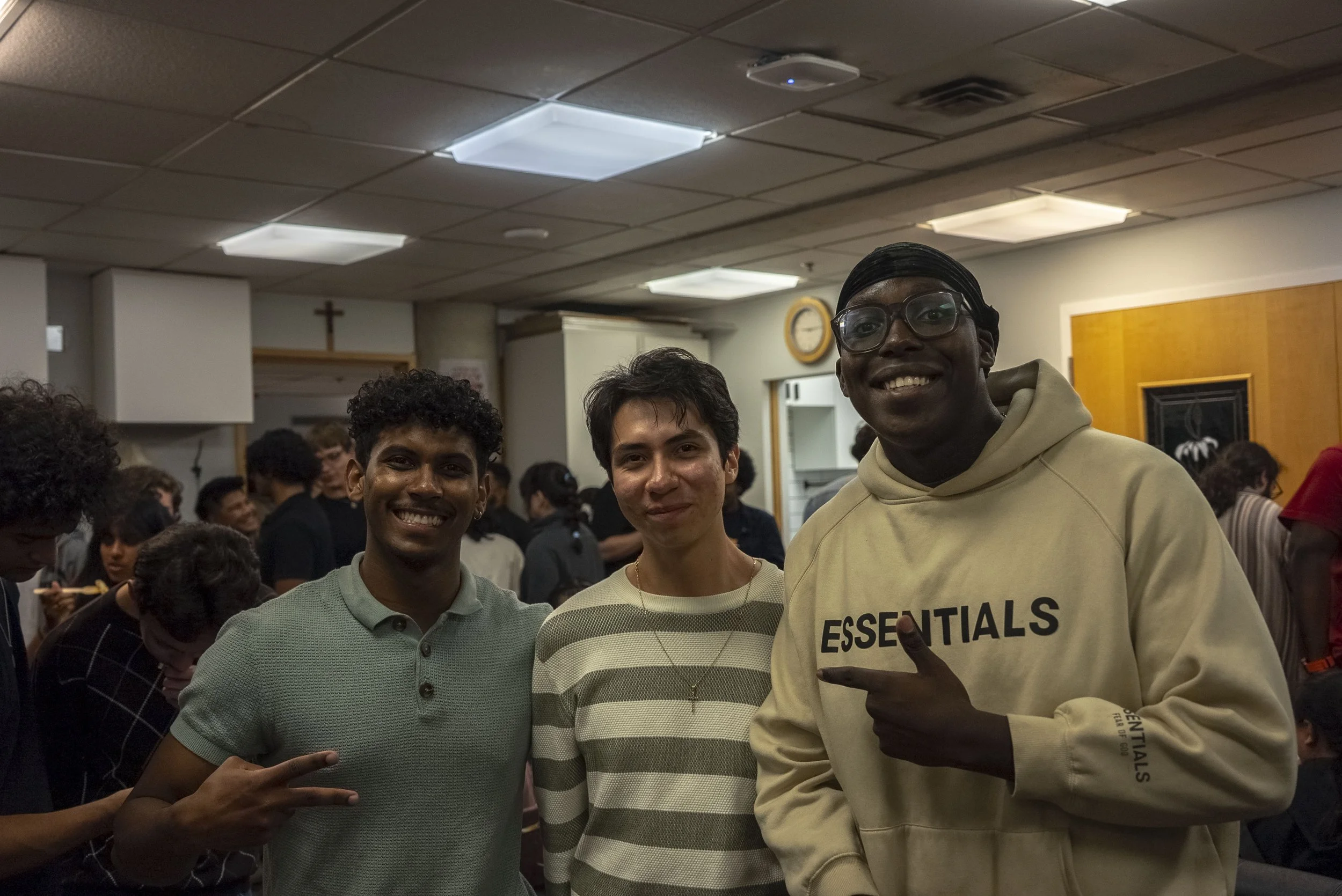 Three young men standing in a crowded room, smiling and posing for the camera. The room has a drop ceiling with rectangular lights, and there is a wooden cabinet and a clock on the wall in the background. The young men are wearing casual clothing, with one in a beige hoodie that says 'ESSENTIALS', and they appear to be happy and enjoying the moment.