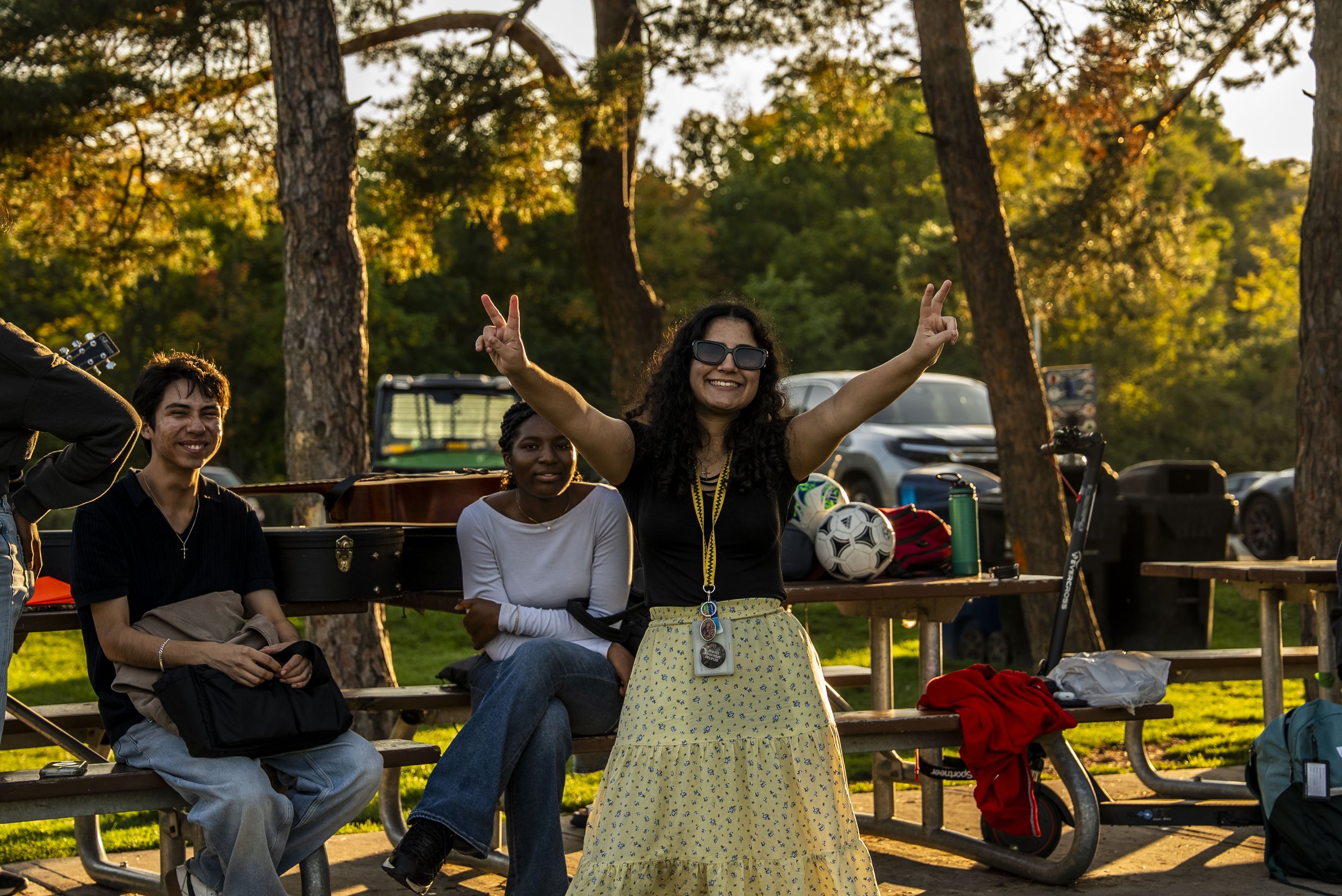 Group of people outdoors in a wooded park during daytime. One woman in the center wearing sunglasses and a yellow dress with a clouds pattern is smiling and posing with peace signs. Two women are seated on benches around her, and other items like a soccer ball, backpacks, and water bottles are visible.