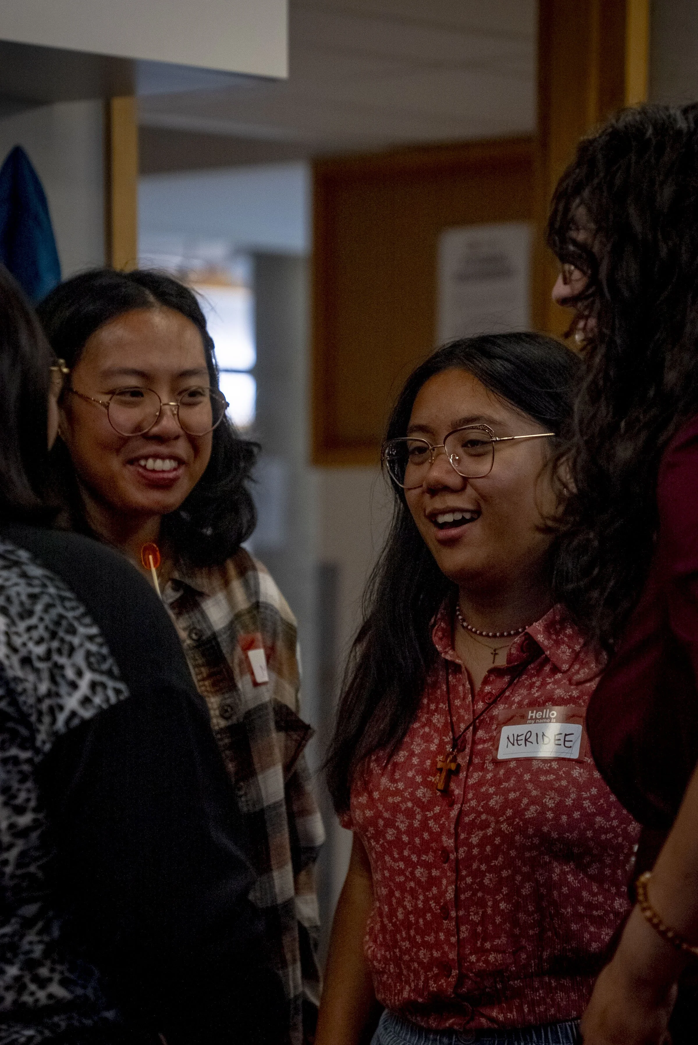 Four women engaged in conversation, one wearing a name tag that says 'NERIDEE,' in an indoor setting with wooden and neutral-colored walls.