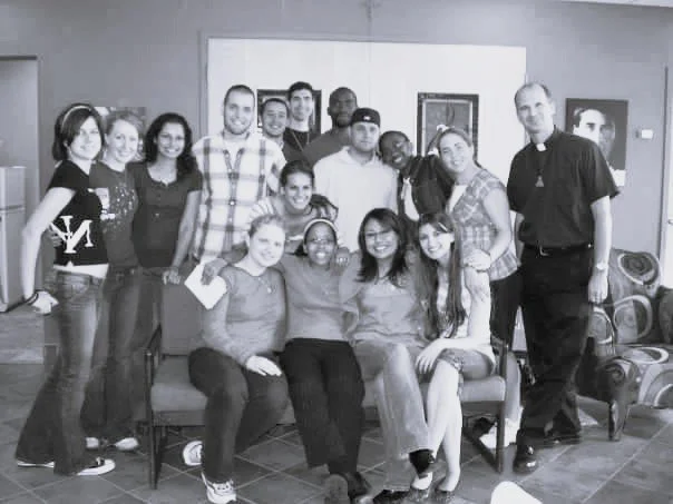 Group of young people and a man, possibly a priest, gathered together in a room, some sitting on a couch and others standing behind, posing for a photo.