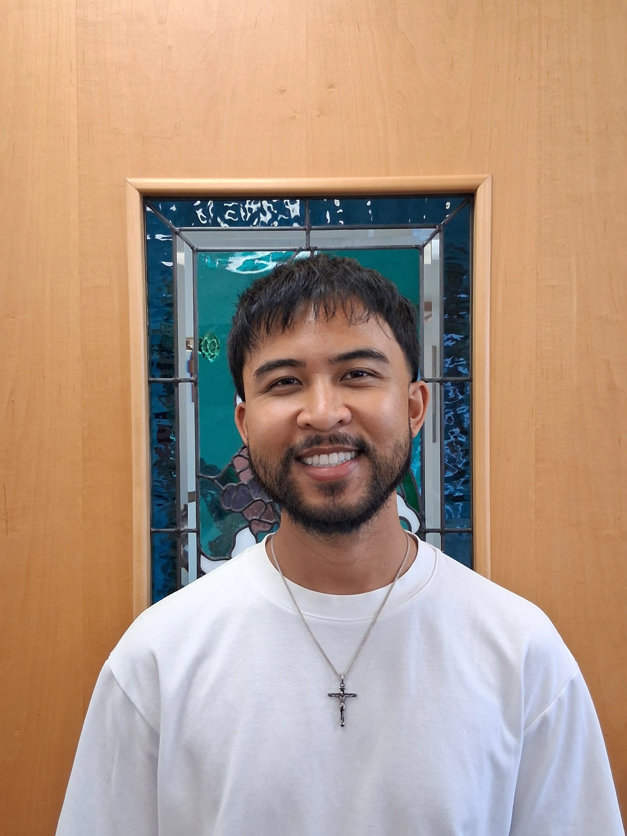 Close-up of a smiling young man with short dark hair and a beard, wearing a white shirt and a silver cross necklace, standing in front of a wooden wall with a decorative mirror behind him.